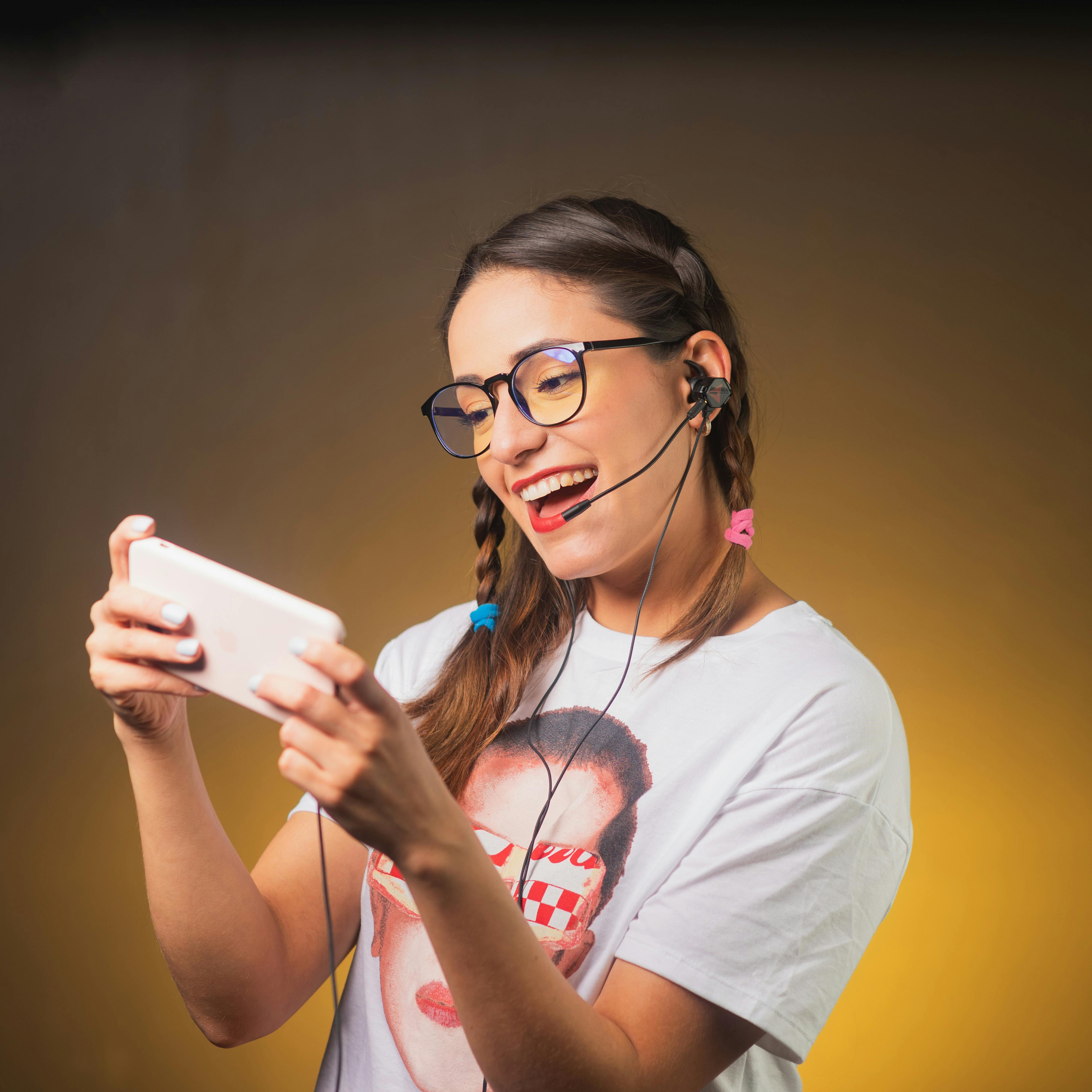 Happy woman with glasses and headset joyously playing games on a smartphone with a bright background.
