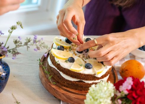 Hands decorating a lemon blueberry cake with herbs, surrounded by fresh flowers.