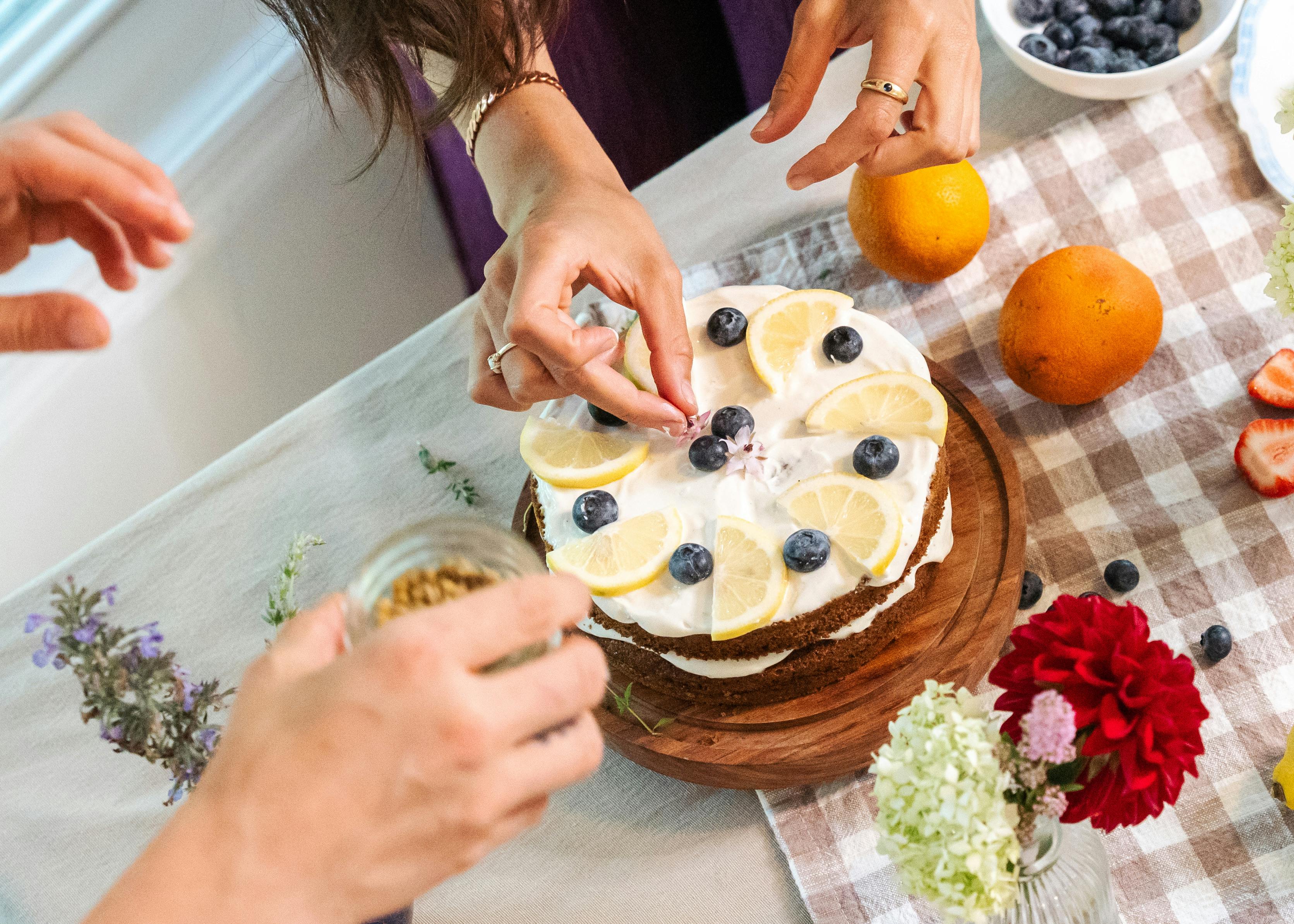 Hands decorating a lemon and blueberry cake with fresh fruits on a summer day.