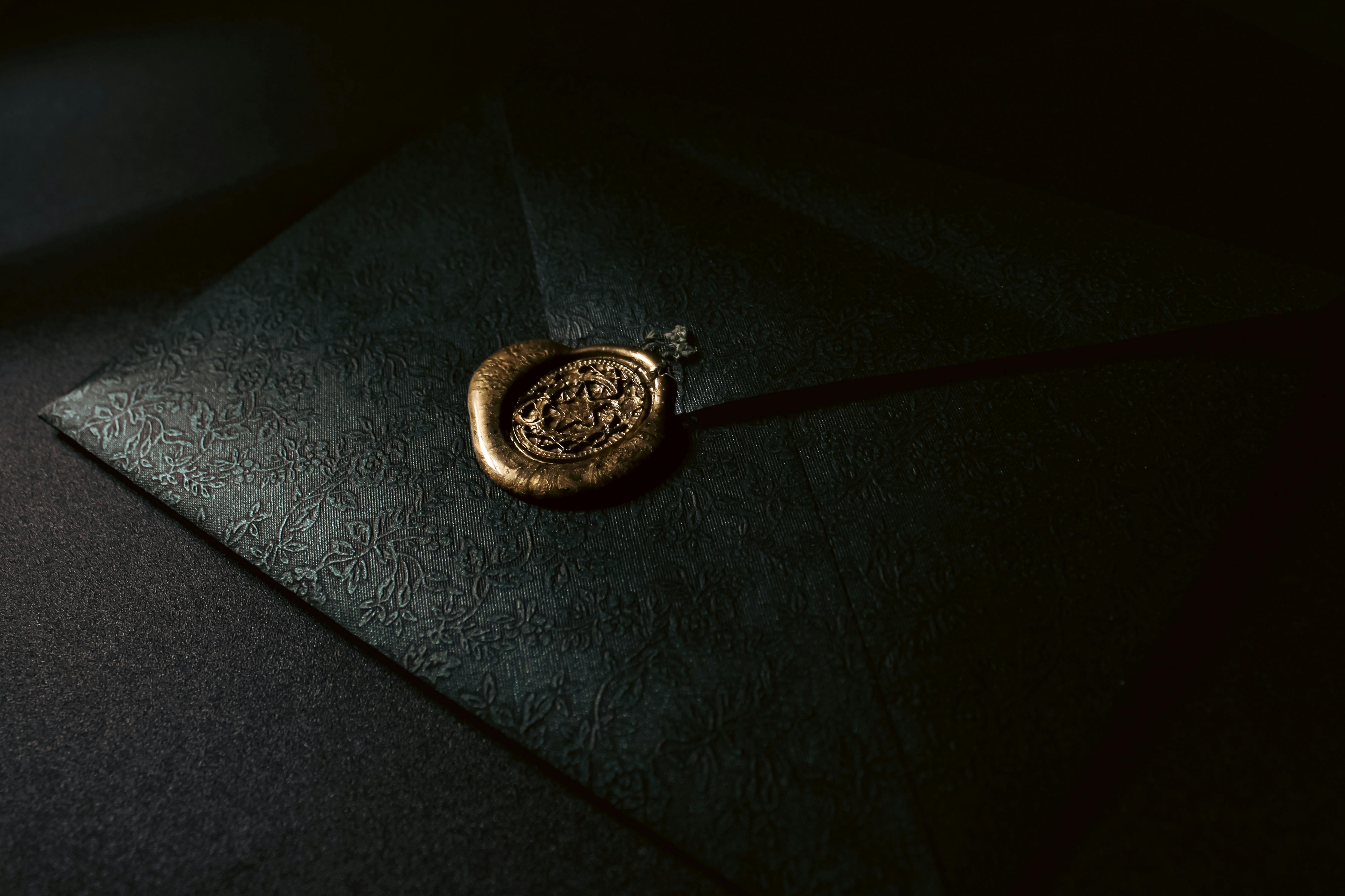 Kostenlos Eleganter schwarzer Umschlag mit geprägtem Muster und goldenem Wachssiegel in stimmungsvoller Lichtatmosphäre. Stock-Foto