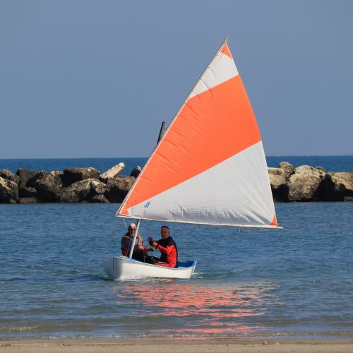 Free Two people enjoy sailing on a small boat with an orange and white sail near a rocky coast. Stock Photo