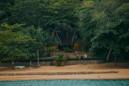 A secluded, thatched-roof house nestled in tropical trees on a sandy beach.