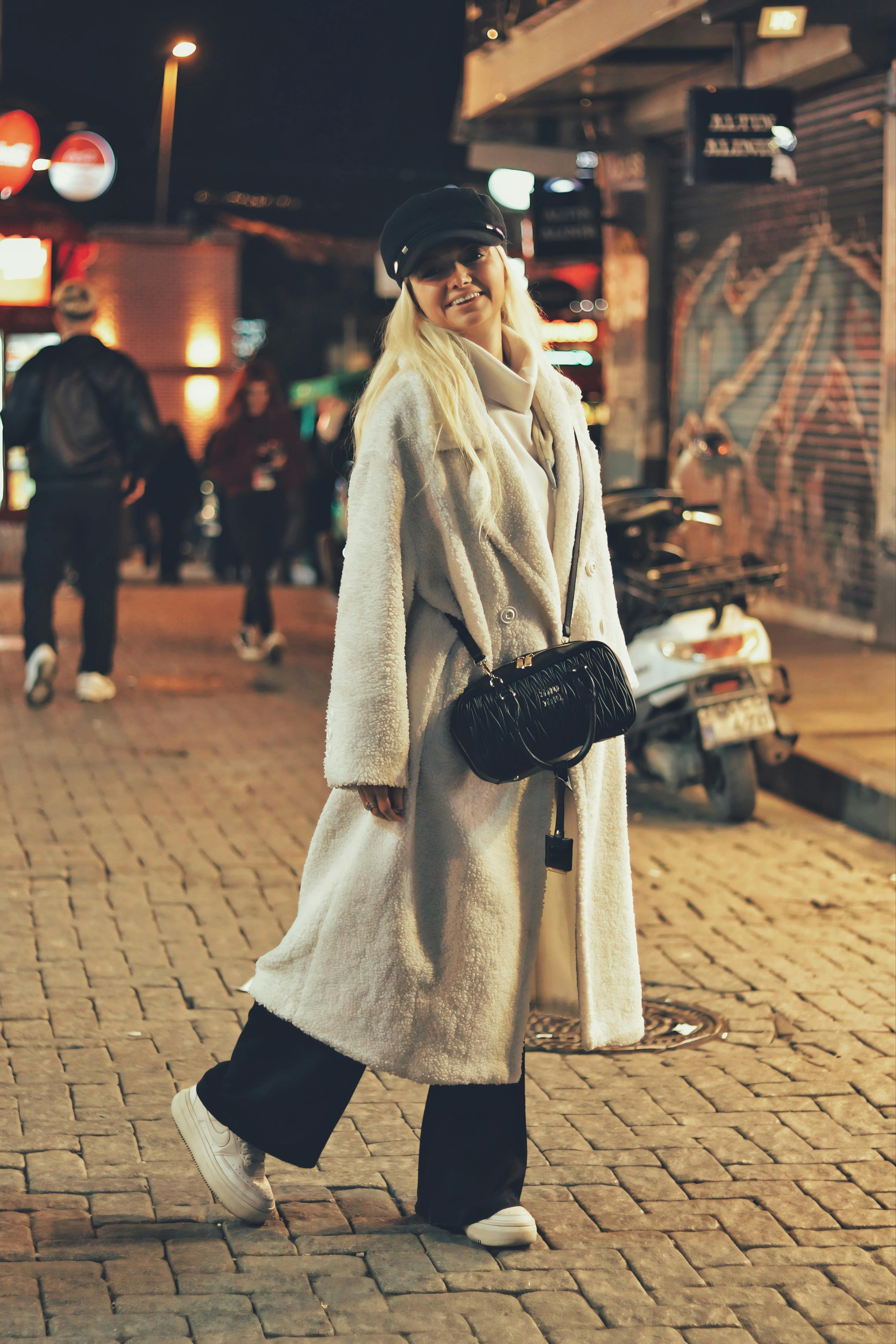 Fashionable woman in a stylish coat and cap walking down a lively city street at night.