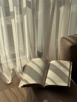 A tranquil reading spot with a book and soft sunlight casting shadows through curtains in İzmir, Türkiye.
