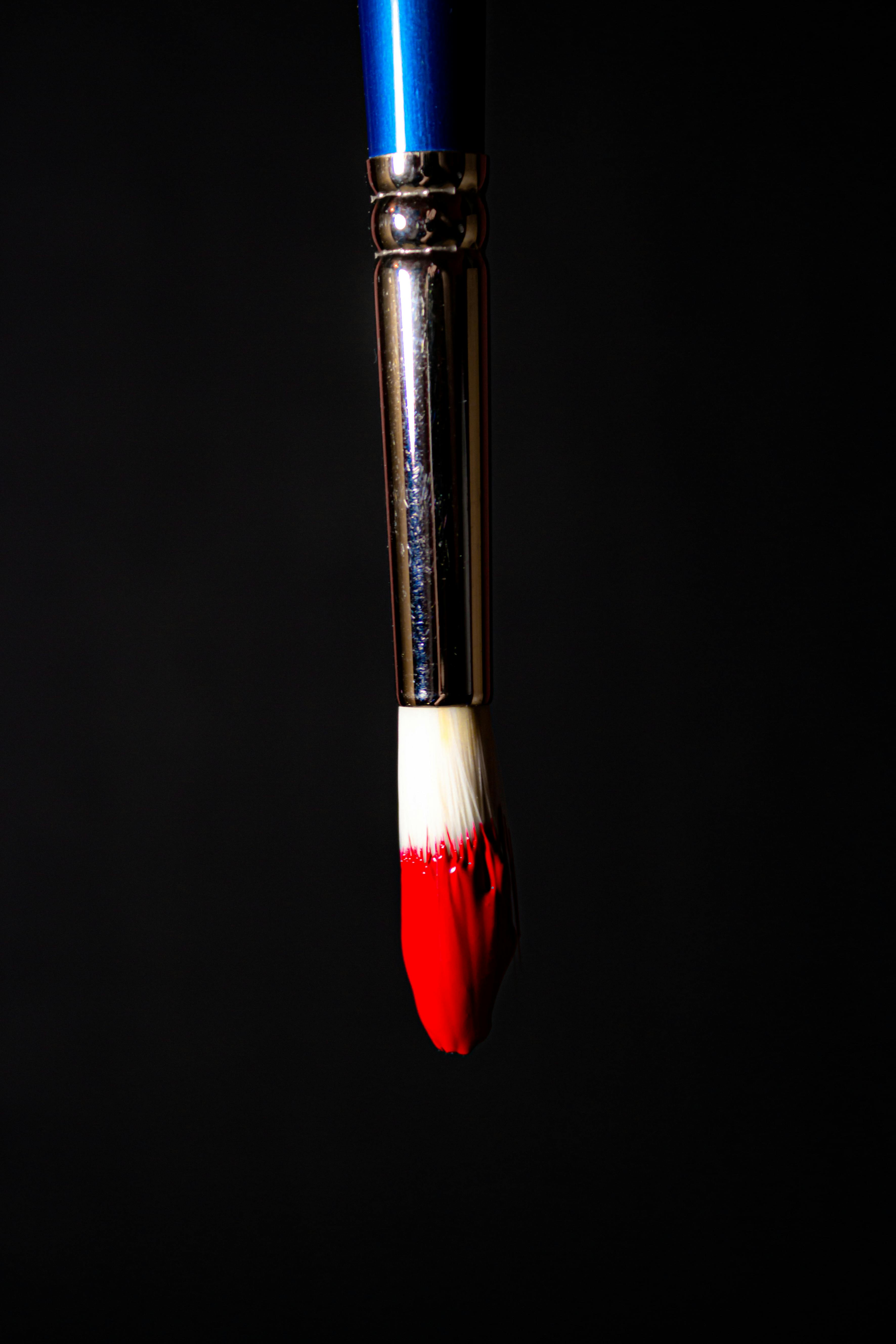 Artistic close-up of a paintbrush dripping vibrant red paint against a dark background.