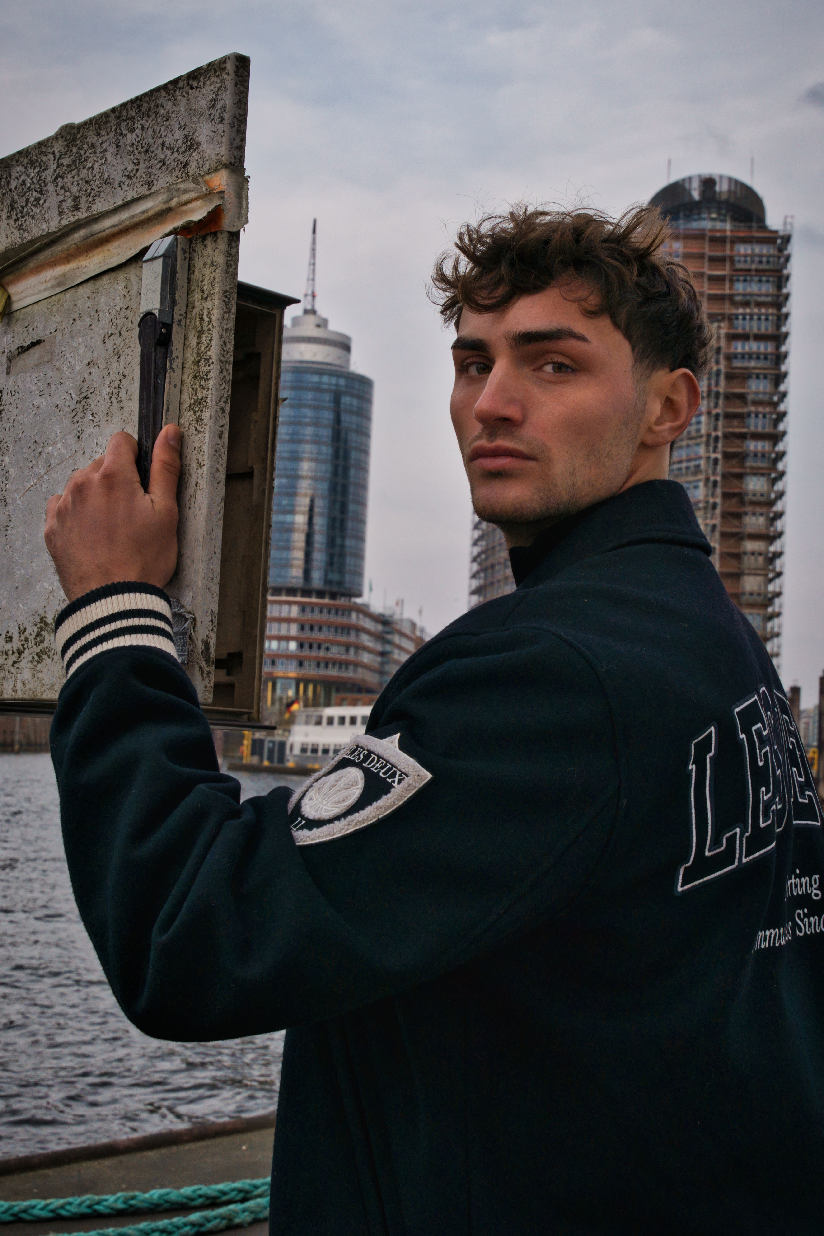 A young man poses by the river with urban architecture in the background, exuding a cool and contemplative vibe.