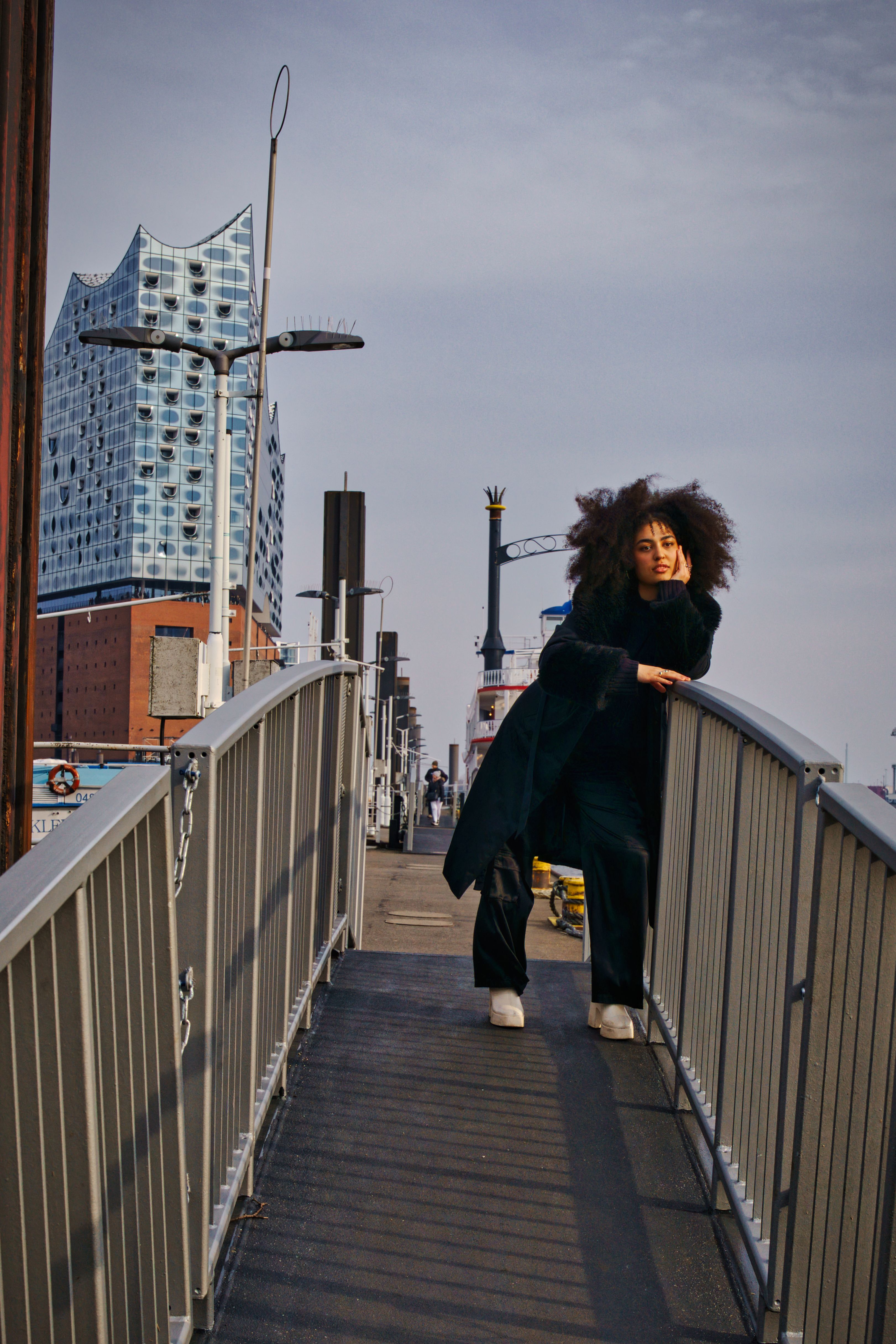 Fashionable woman posing on a bridge with the Elbphilharmonie Hamburg in the background.