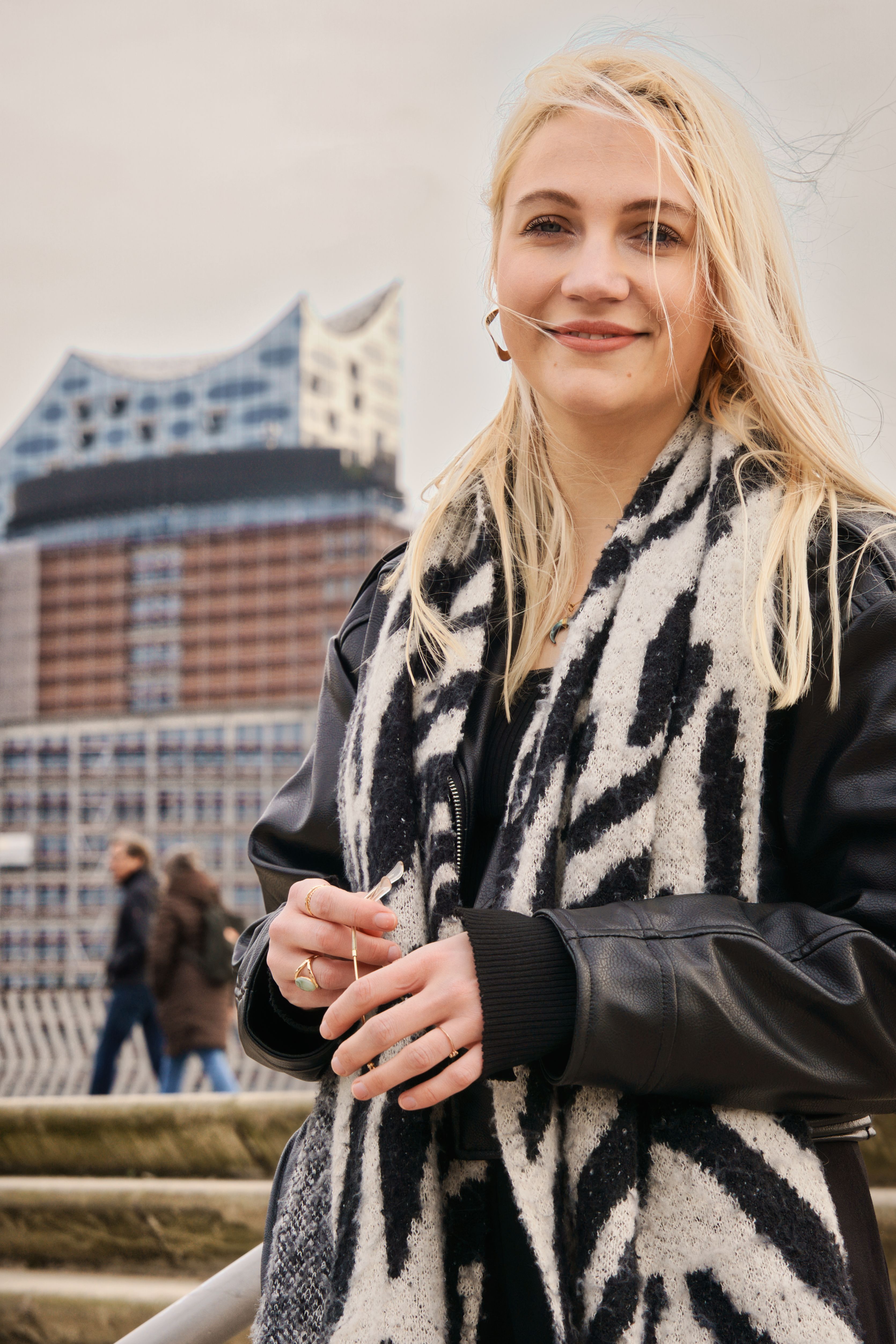 Smiling woman standing outdoors near the Elbphilharmonie in Hamburg, Germany on a windy day.
