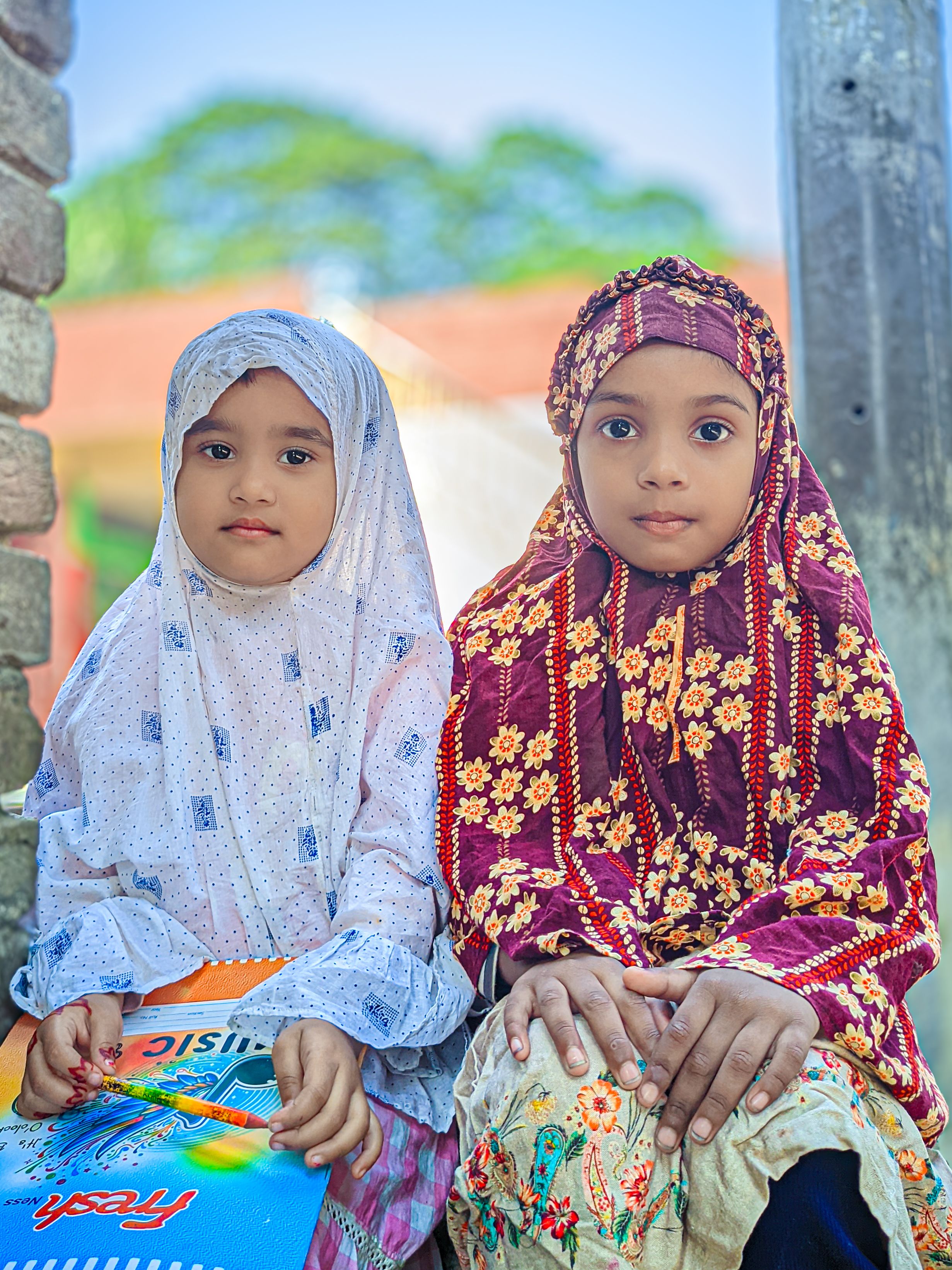 Two young girls wearing traditional clothing, sitting outdoors with books.