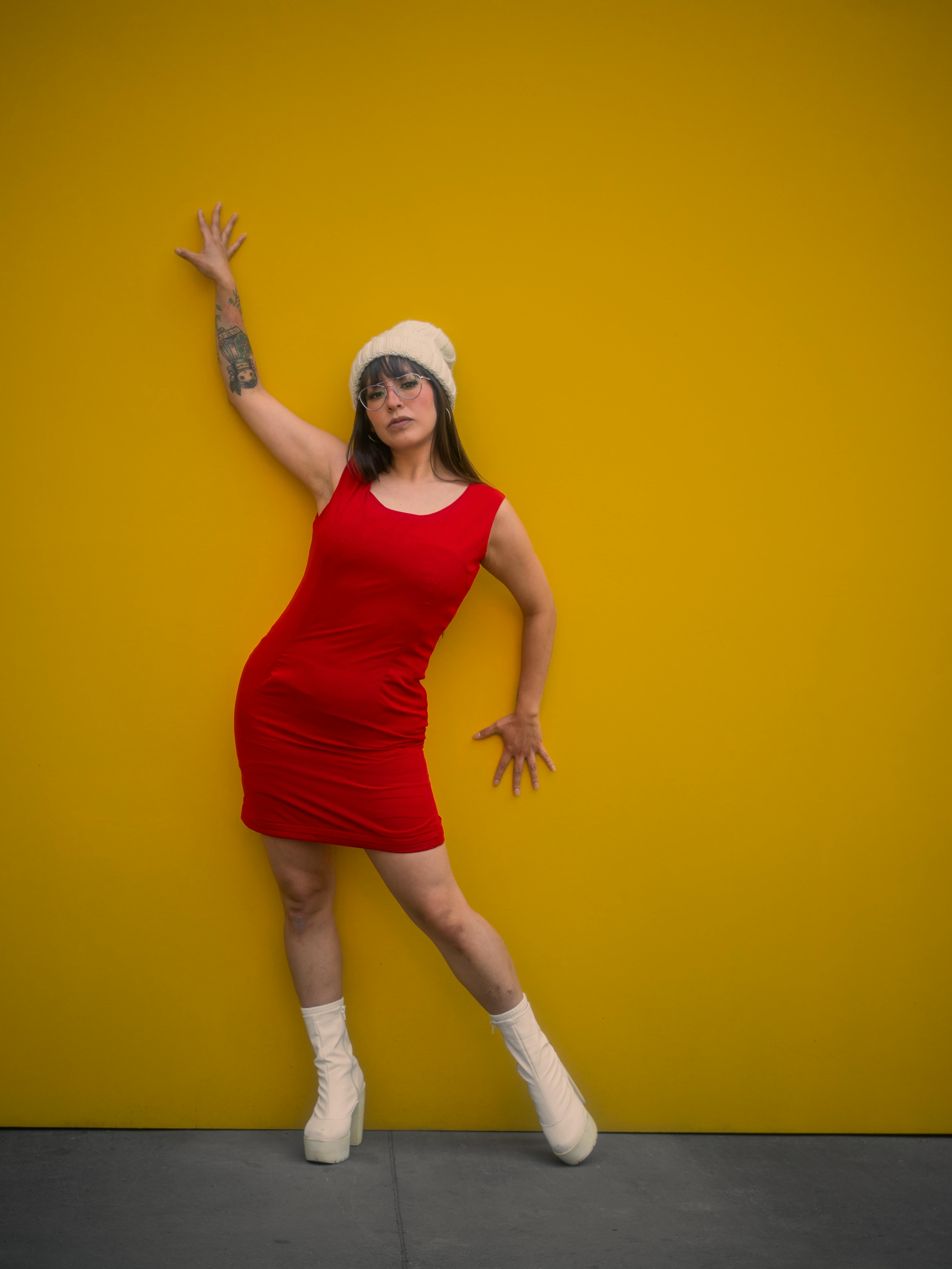 Gratis Una donna elegante posa con un abito rosso e un cappello su uno sfondo giallo brillante. Foto a disposizione