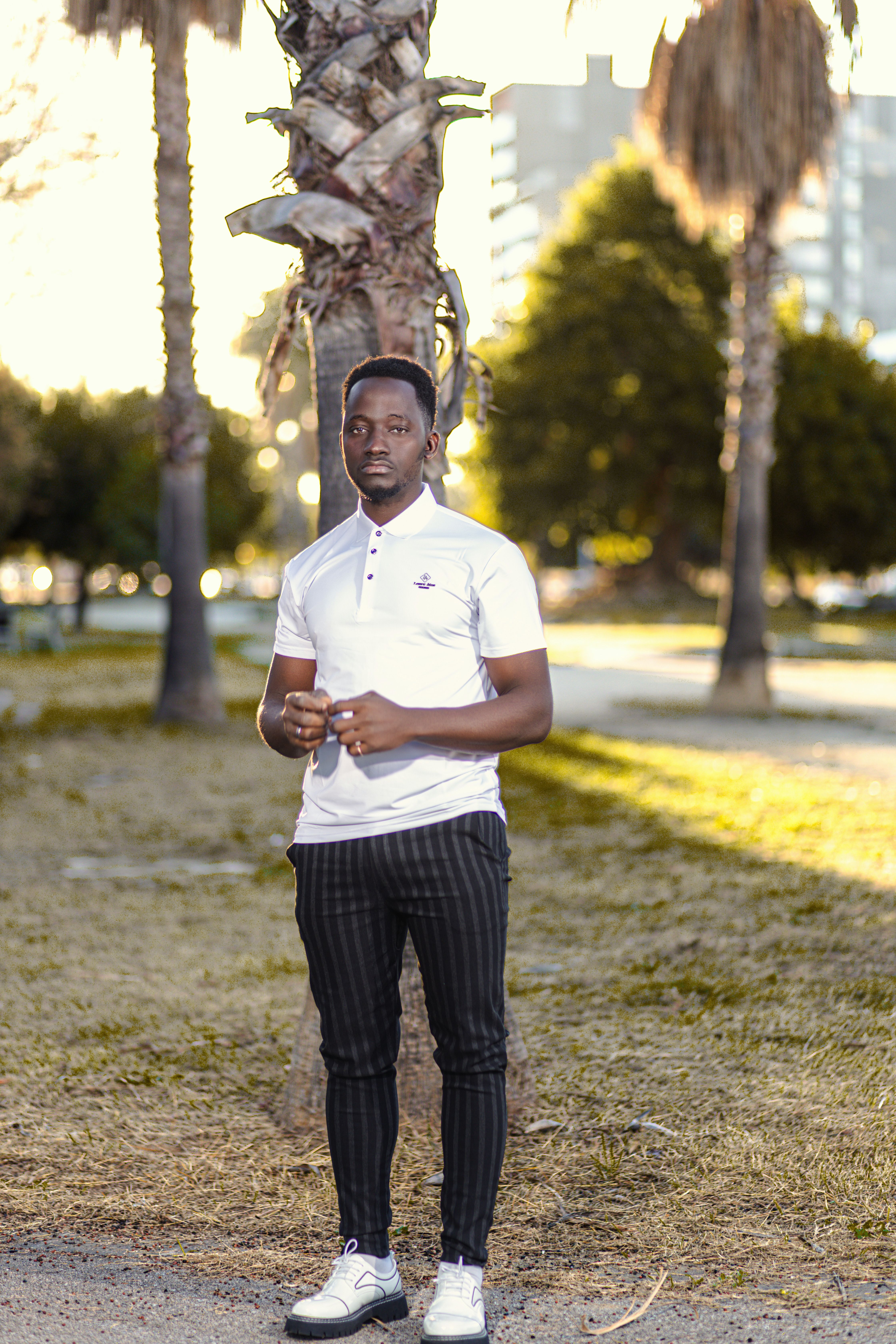 Young man in white polo standing near palm trees in a city park setting.