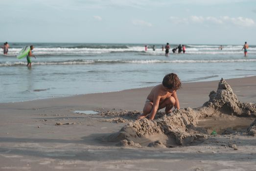 Full body of small boy in panties building sandcastle while resting on beach of ocean in sunlight