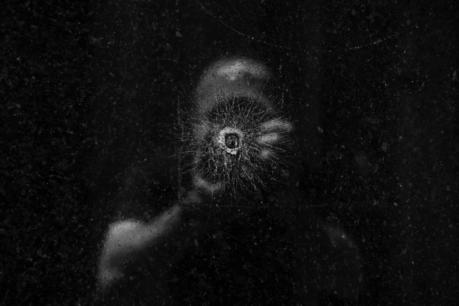 Artistic photograph of a person's reflection seen through a bullet hole on a dark glass surface.