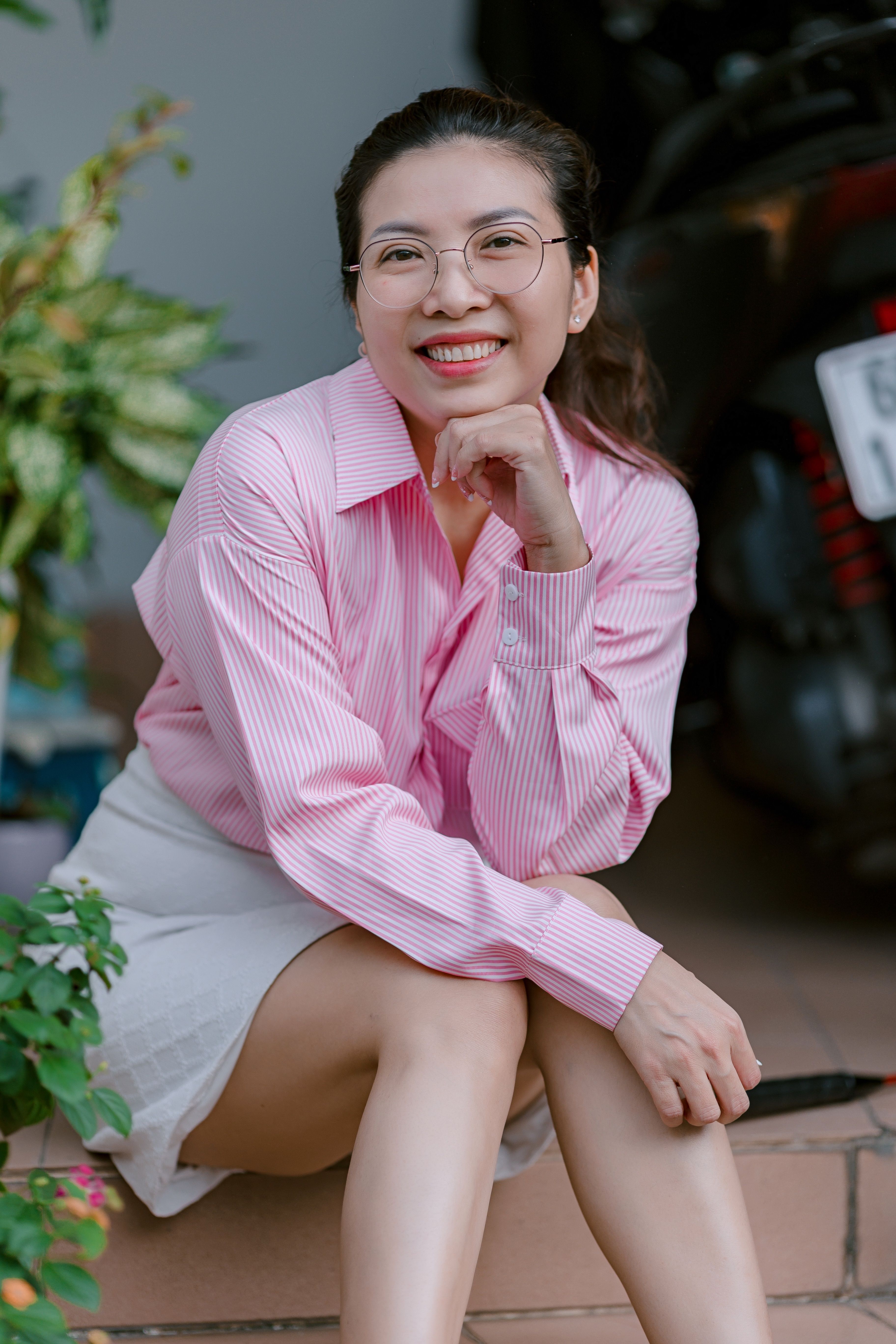 Cheerful woman in pink shirt and glasses sitting outdoors with a warm smile.