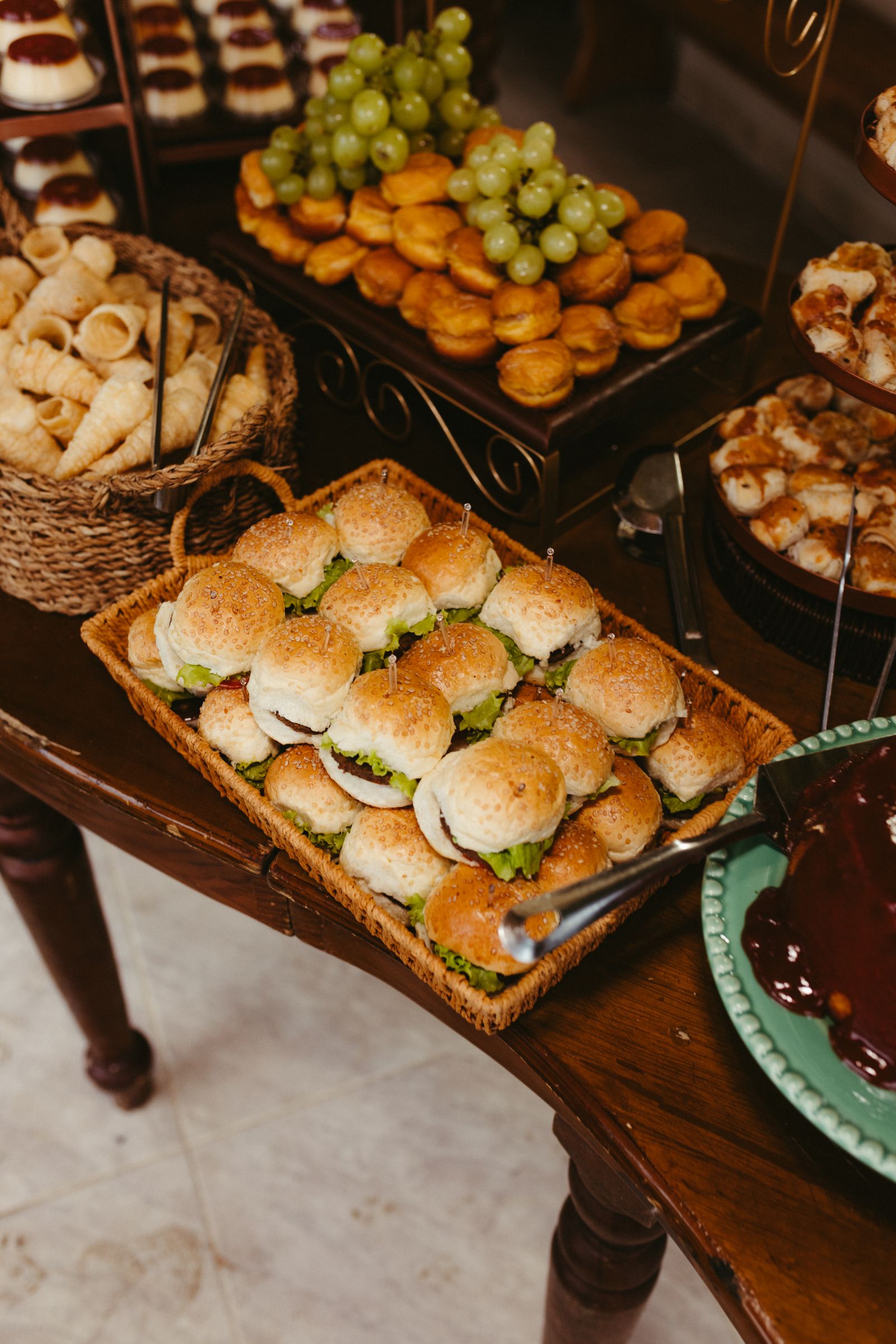 Buffet tray of mini sandwiches beside pastries, grapes, and small desserts.