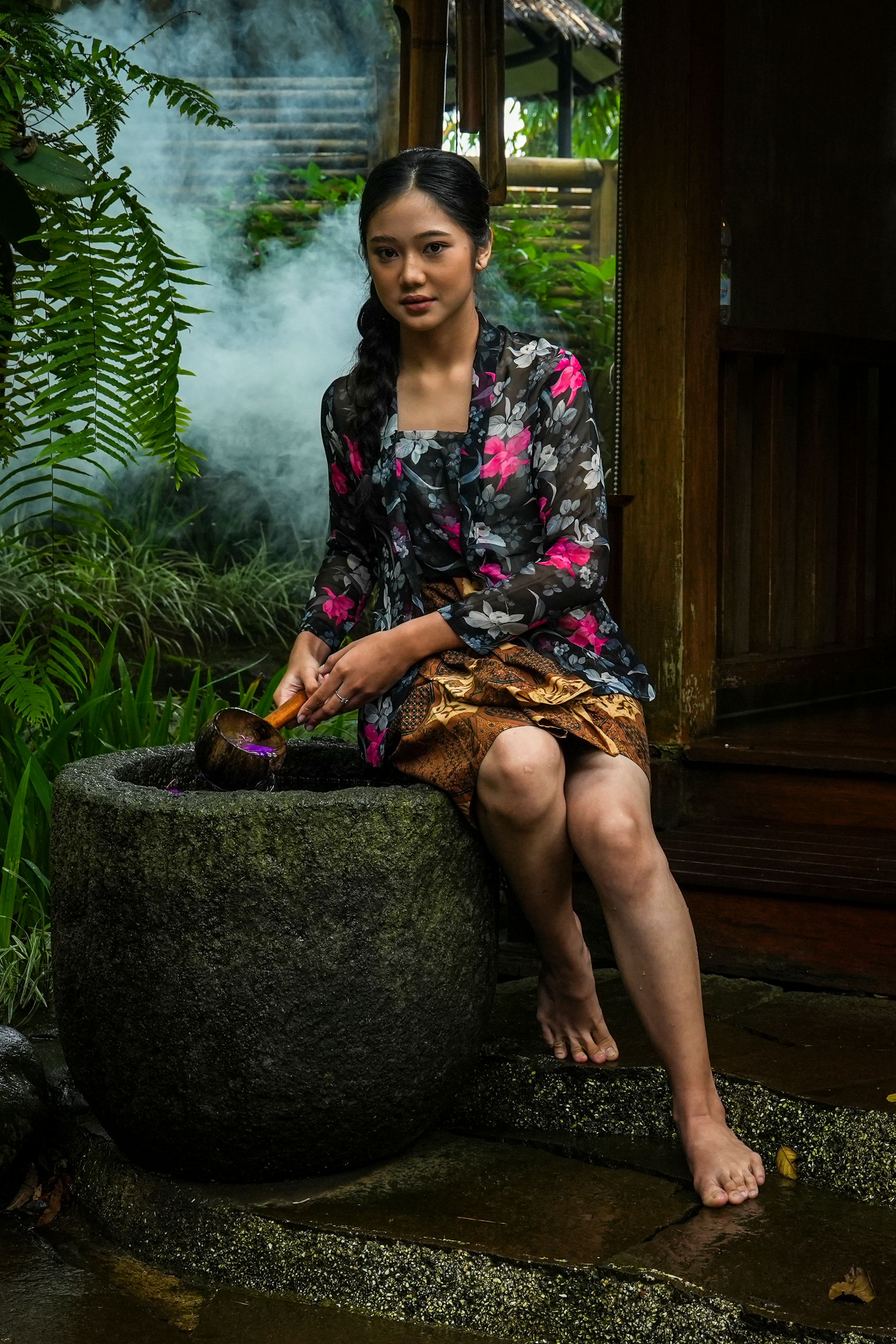 Young Indonesian woman in traditional attire sits outdoors by a stone pot.
