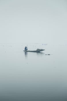 A lone fisherman rows a boat on a foggy lake, creating a serene and minimalist atmosphere.