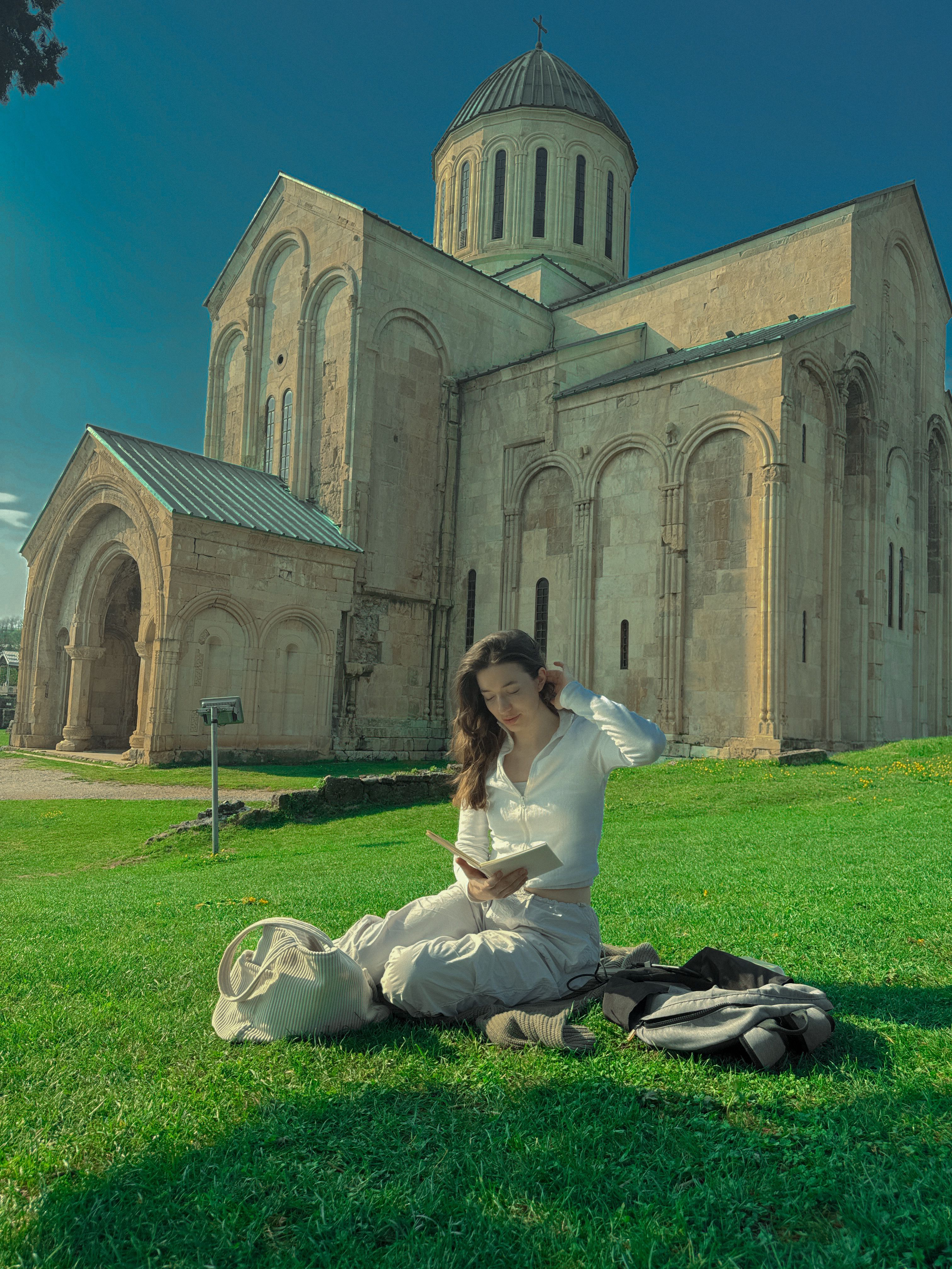Woman reading on grass near the historic Bagrati Cathedral in sunny weather.