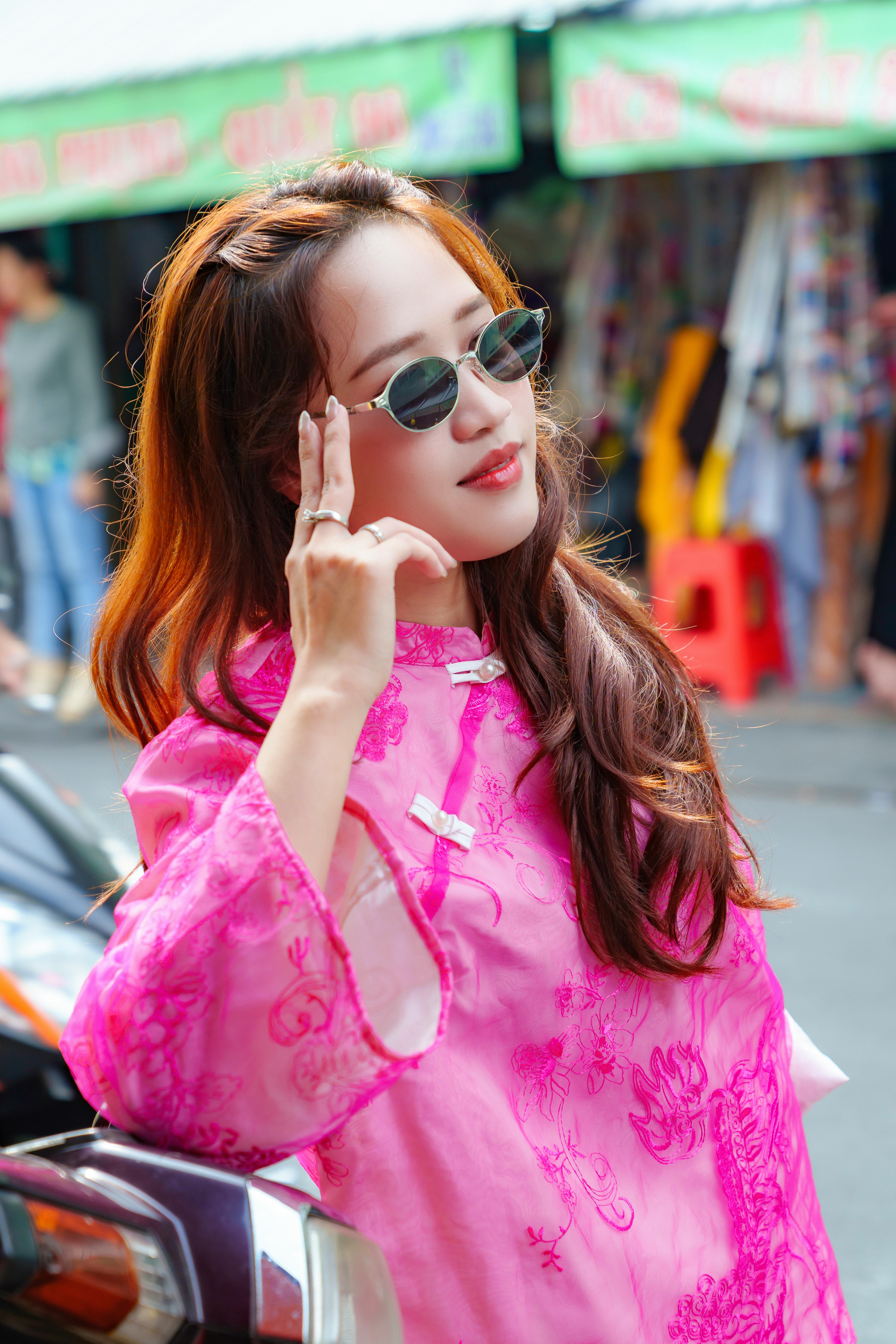 Stylish woman wearing a pink ao dai and sunglasses poses confidently in a vibrant Hồ Chí Minh City market.