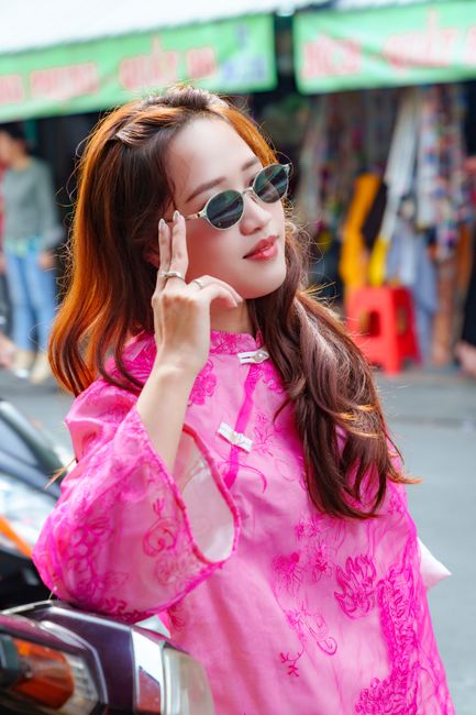 Stylish woman wearing a pink ao dai and sunglasses poses confidently in a vibrant Hồ Chí Minh City market.