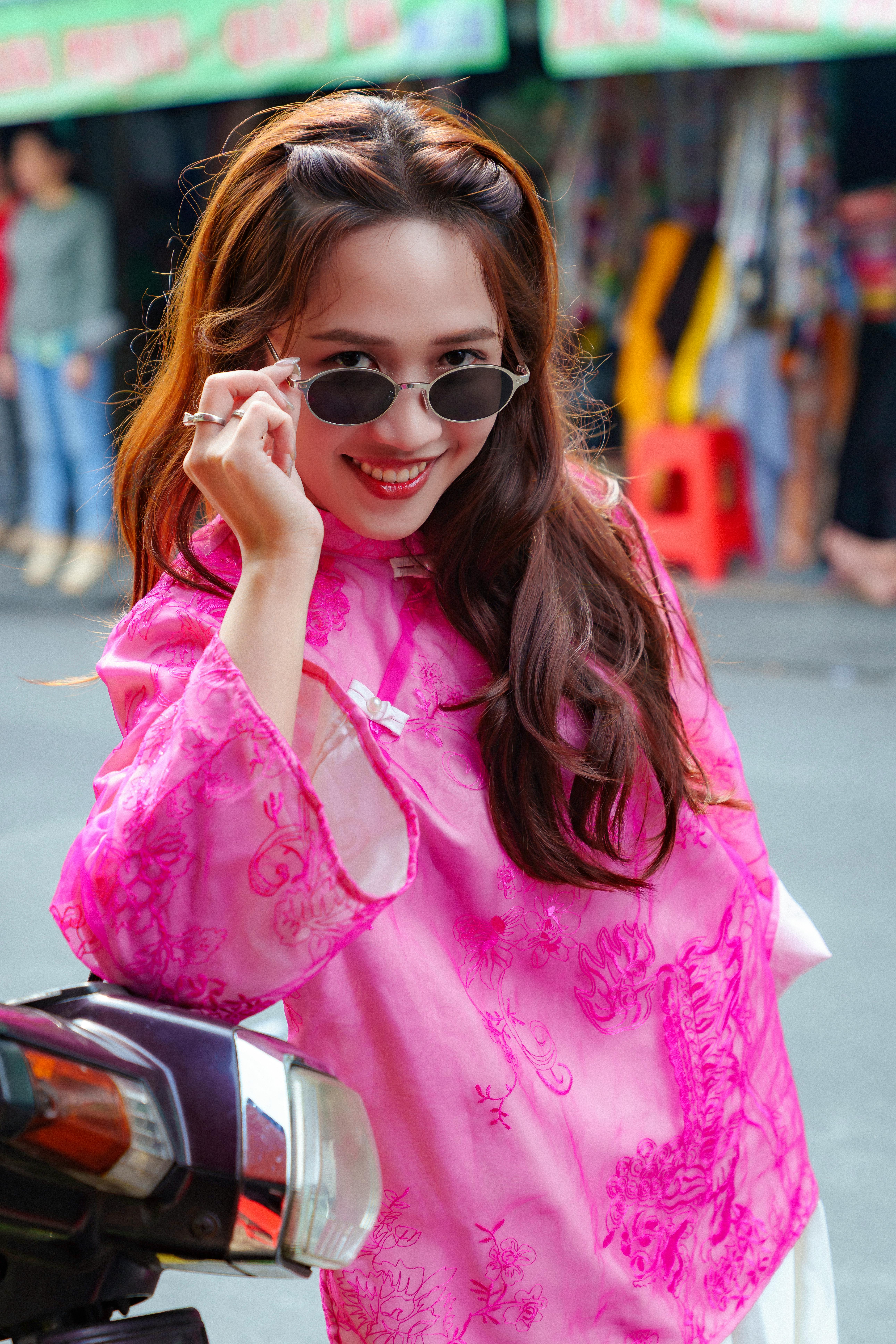 Smiling woman in pink áo dài adjusting sunglasses, on a vibrant street.