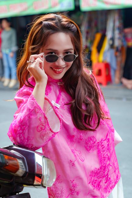 Smiling woman in pink áo dài adjusting sunglasses, on a vibrant street.