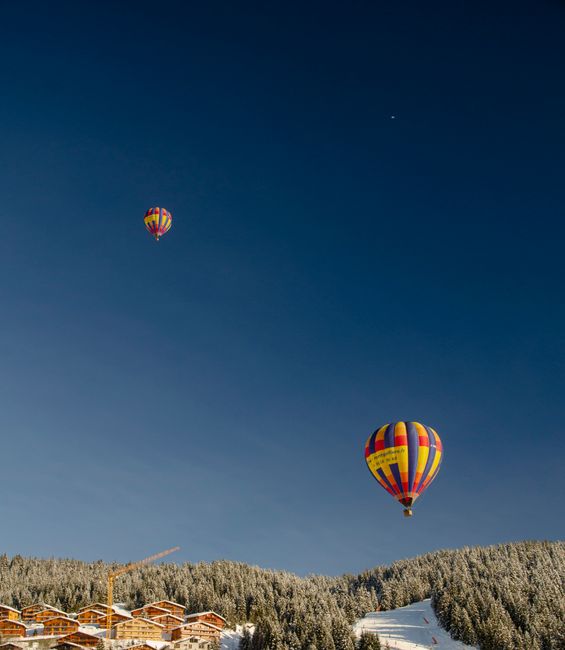 Colorful hot air balloons soaring above a snowy forest and chalet rooftops.