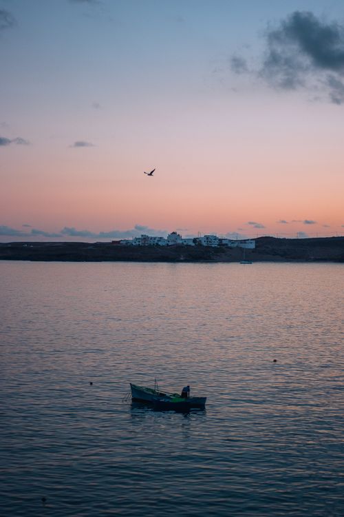 Free A serene sunset scene with a fishing boat on calm waters near a coastal village. Stock Photo