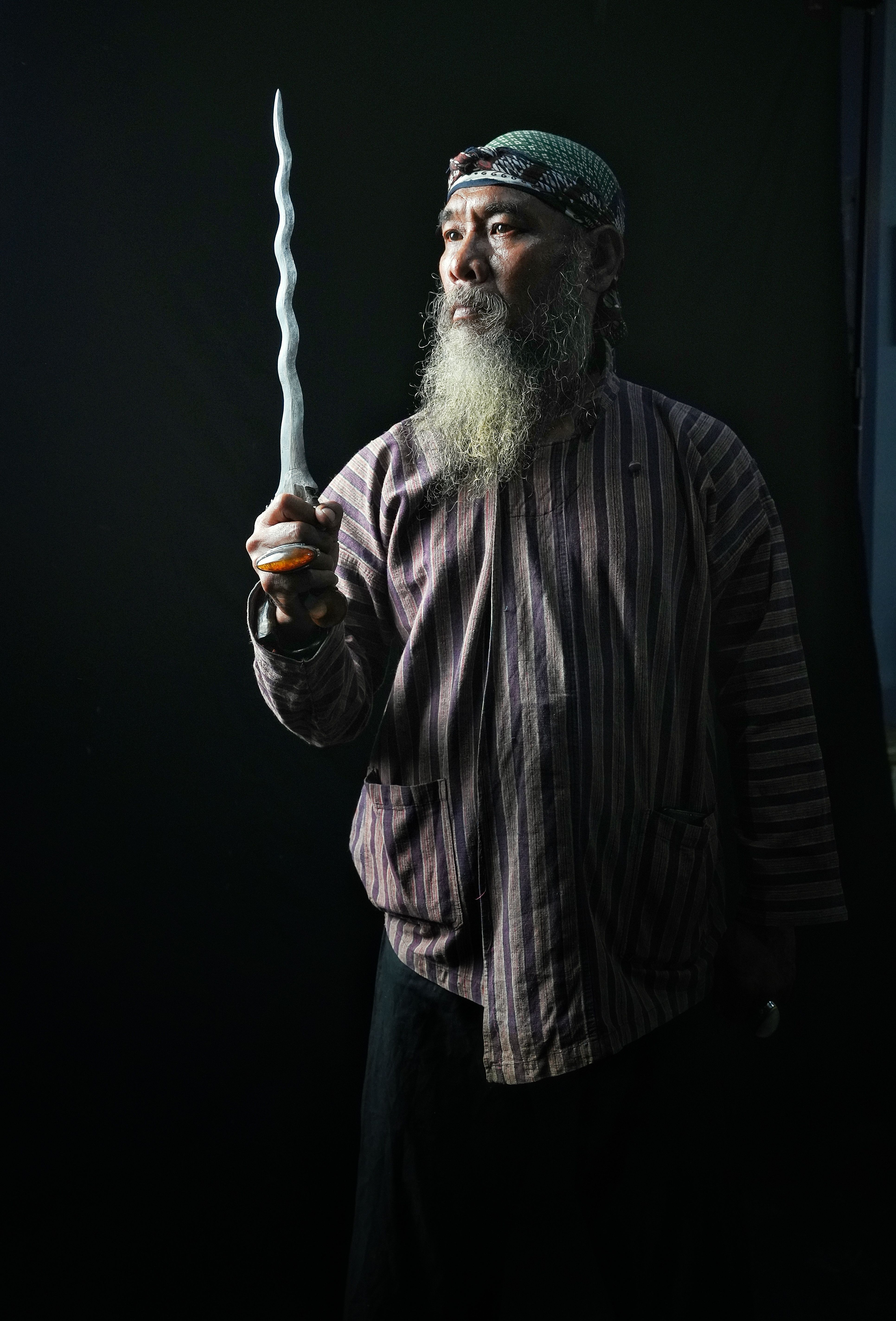 An elderly man in traditional attire holding a spiraled kris dagger against a dark background.