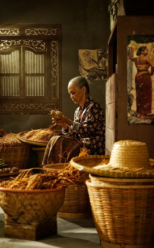 Free An elderly woman sits indoors sorting spices in a traditional setting, surrounded by woven baskets. Stock Photo
