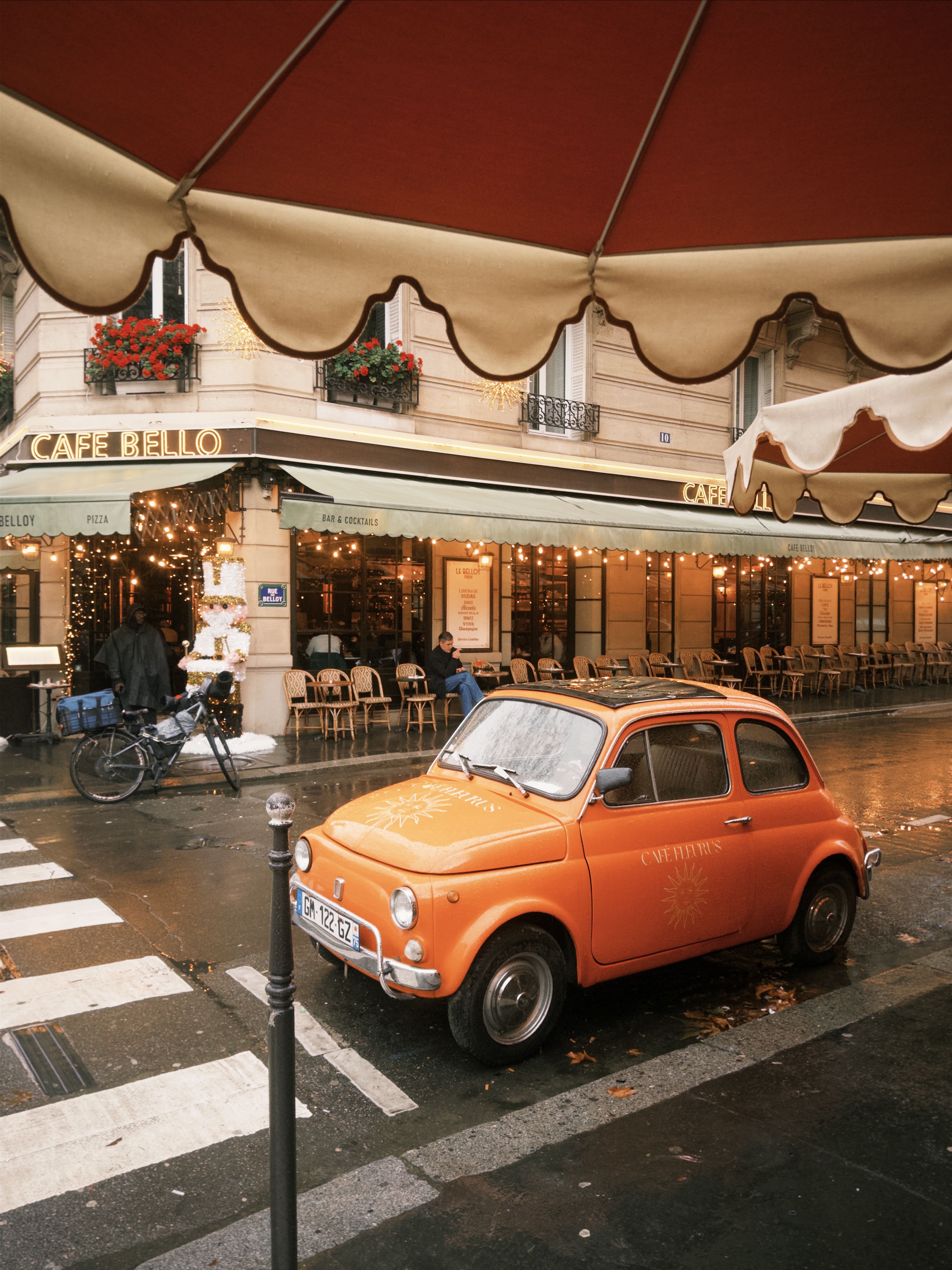 A classic orange car parked outside a charming Parisian cafe on a rainy day.
