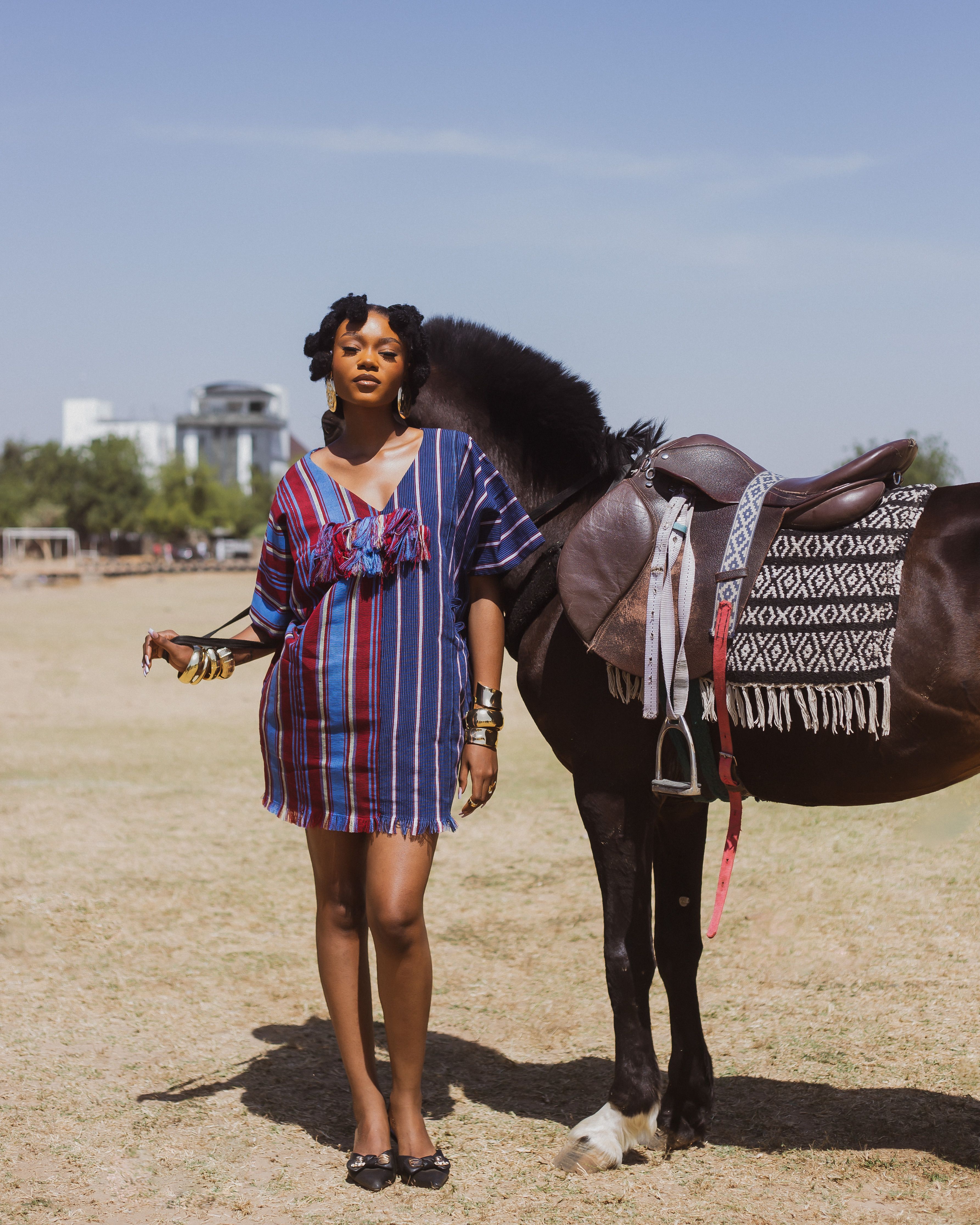 Fashionable woman in striped dress standing beside a horse on a sunny day.