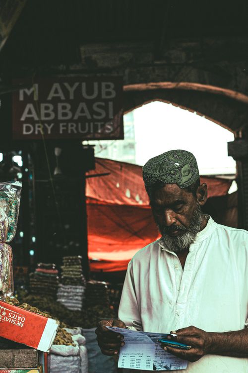Free Elderly man in traditional attire reading a document in a bustling dry fruits market. Stock Photo