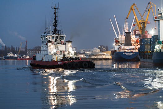 A tugboat navigates a busy port at night, surrounded by cargo ships and cranes.