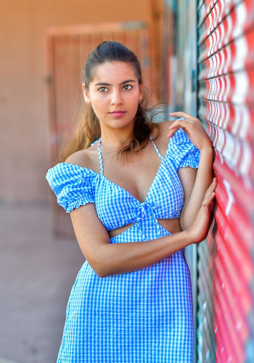 Free Portrait of a young woman in a blue gingham dress leaning against a wall outdoors. Stock Photo