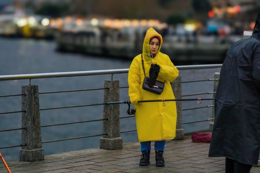 A woman in a yellow raincoat with a fishing rod by a waterfront on a cloudy day.