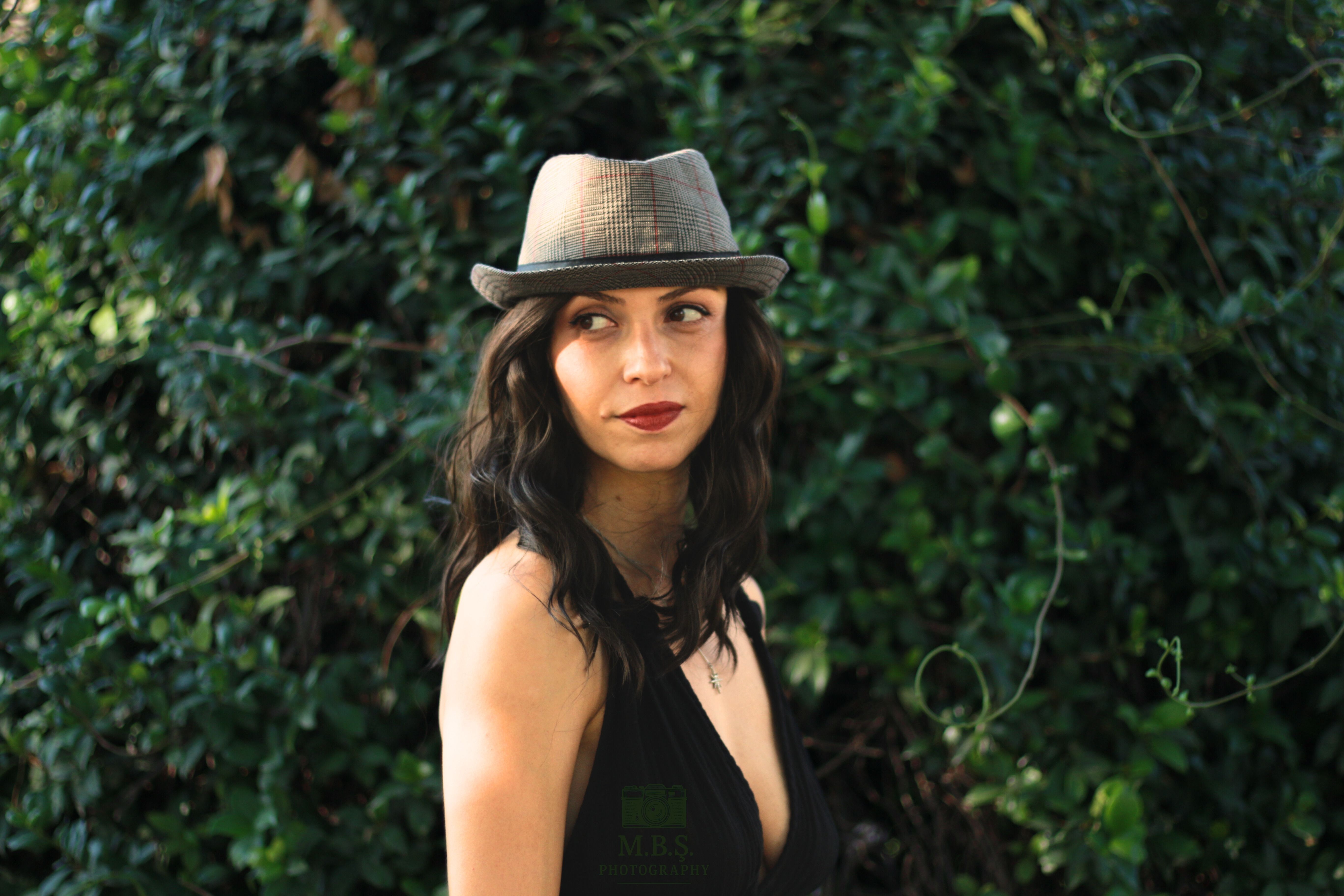 Elegant woman in hat posing outdoors against lush greenery, exuding confidence and style.