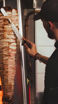 A chef skillfully slices doner kebab meat from a vertical rotisserie in a restaurant kitchen.