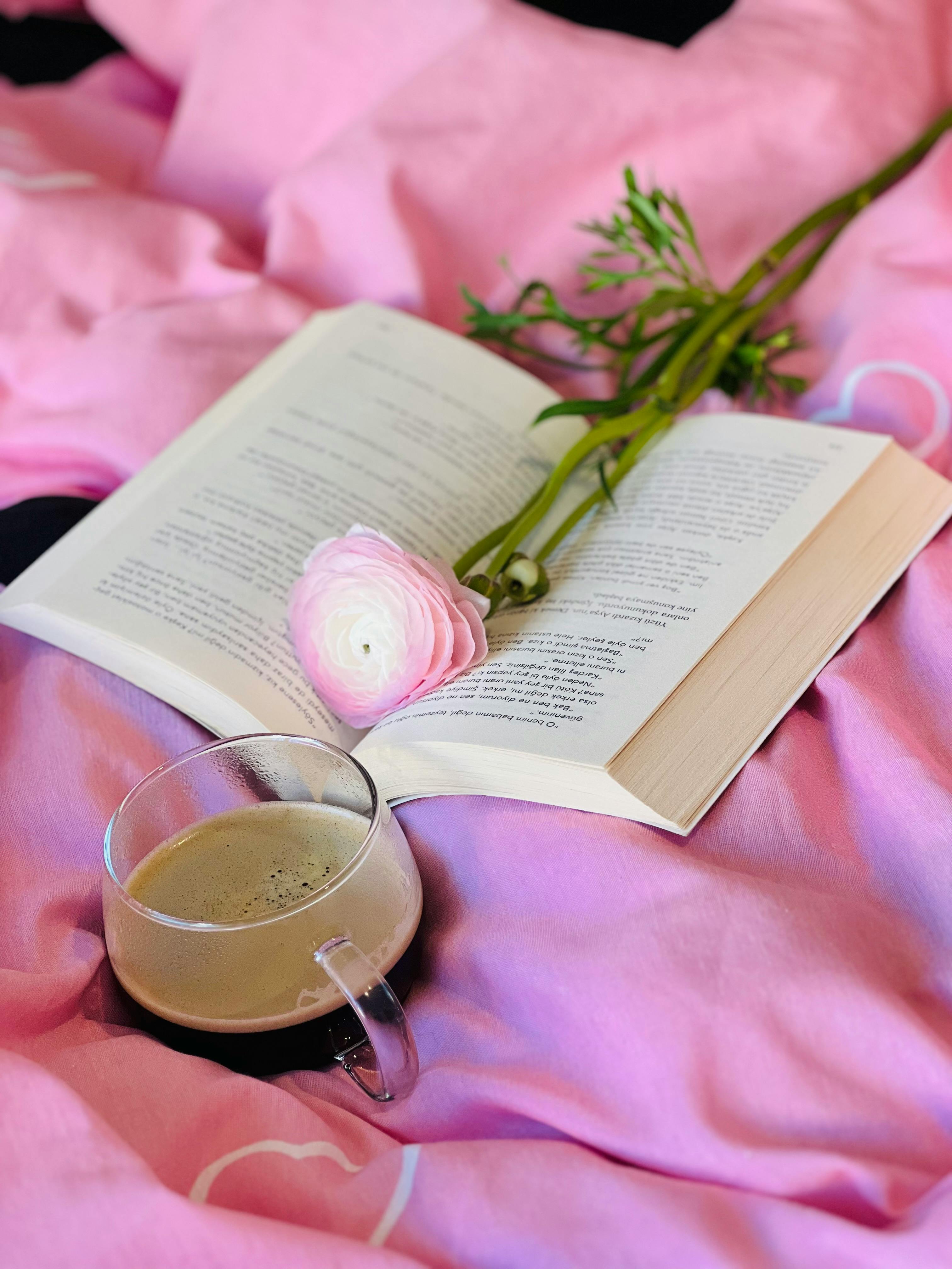Open book with a coffee cup and flower on pink blanket, creating a cozy and peaceful vibe.