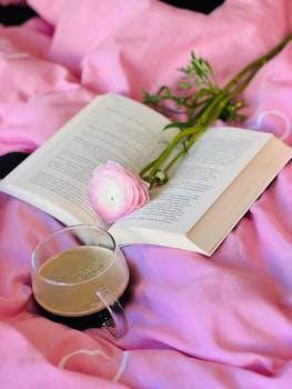 Open book with a coffee cup and flower on pink blanket, creating a cozy and peaceful vibe.