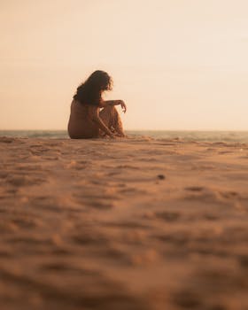 A contemplative woman sits on the sandy beach during a serene sunset, creating a peaceful silhouette.