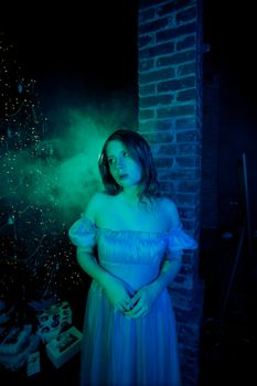 Woman in a blue gown illuminated by blue-green light, beside a Christmas tree with mist.
