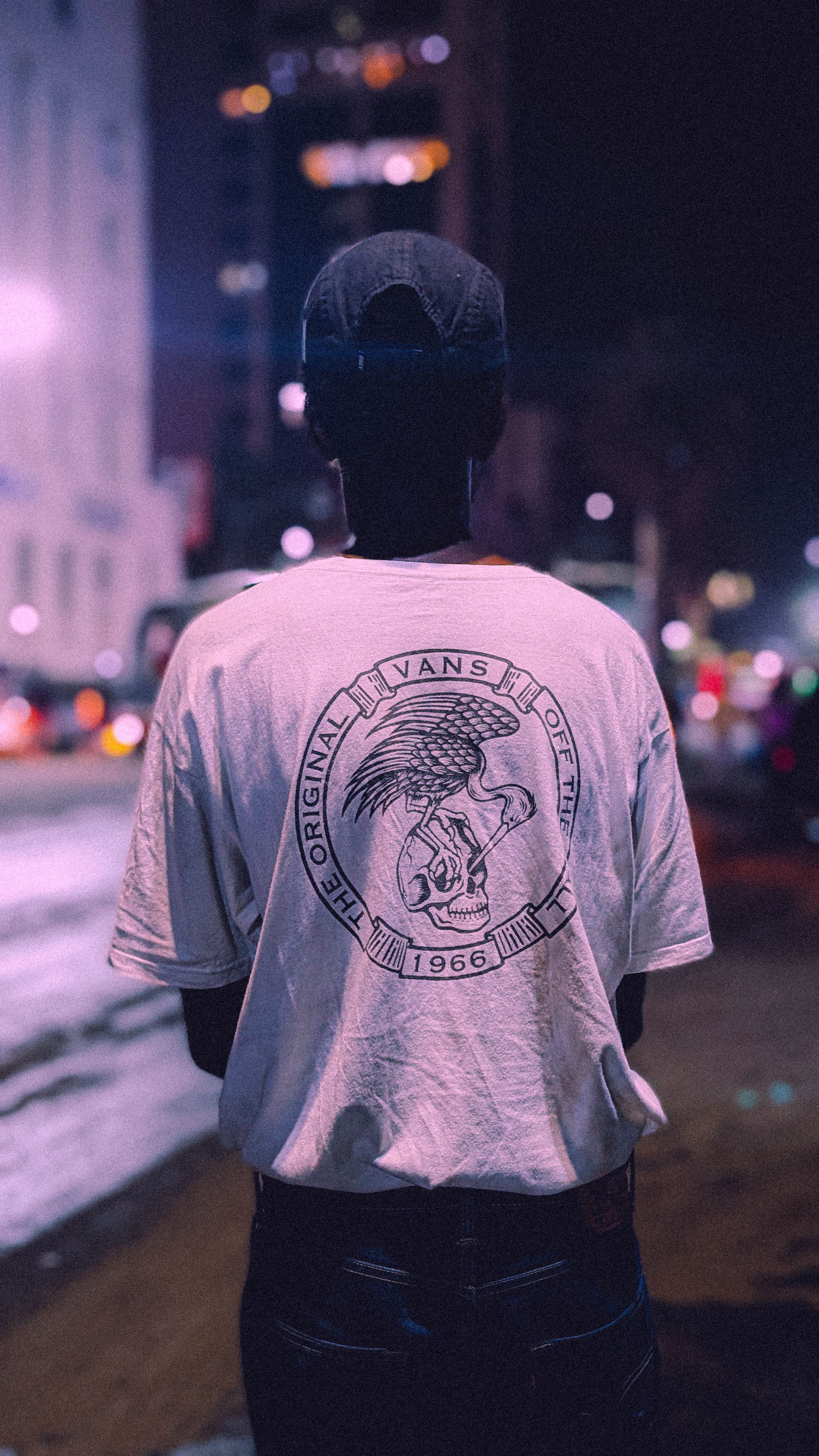 A person stands on a city street at night, wearing a hat and graphic tee.