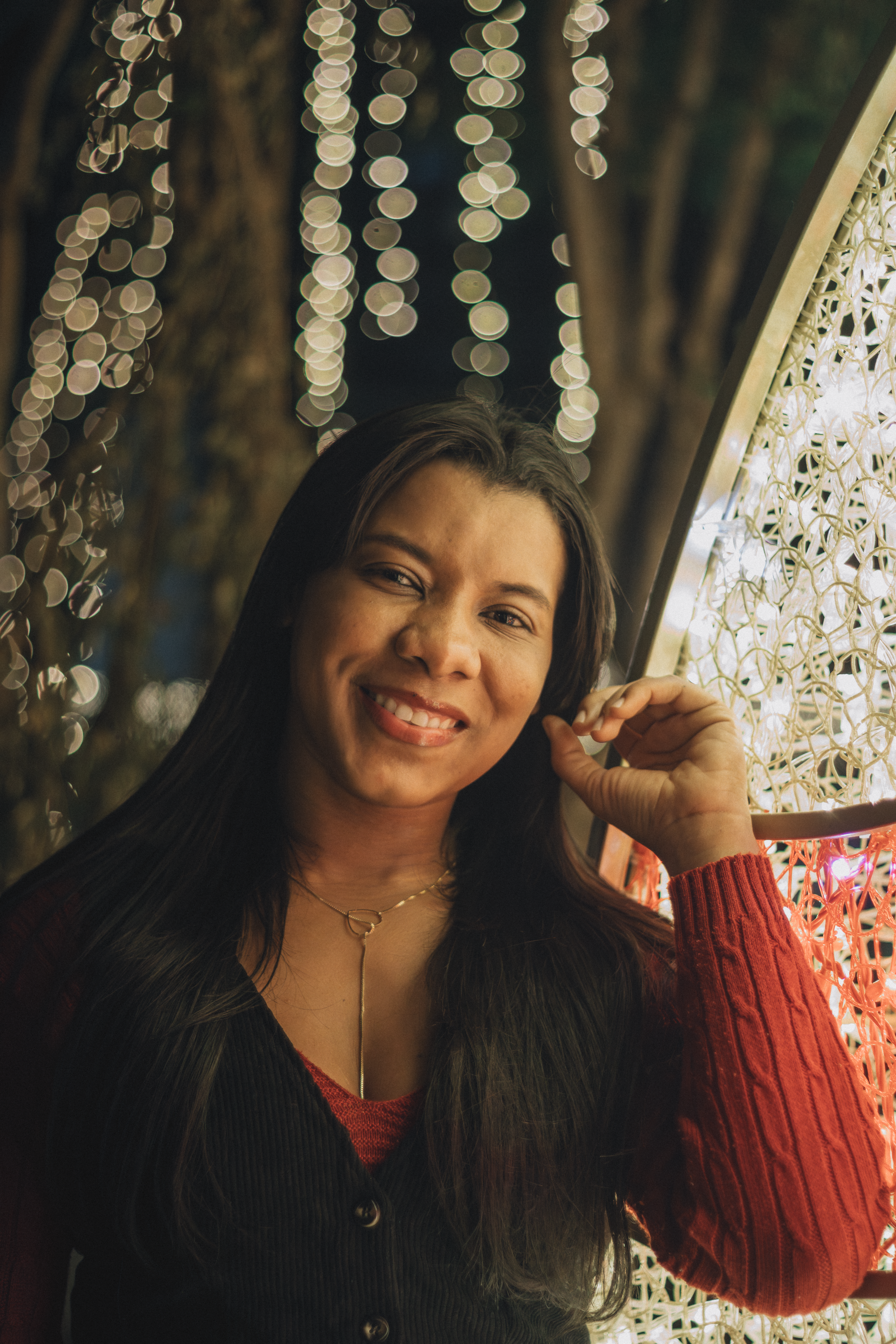 A cheerful woman in a red sweater smiles with festive lights in the background.