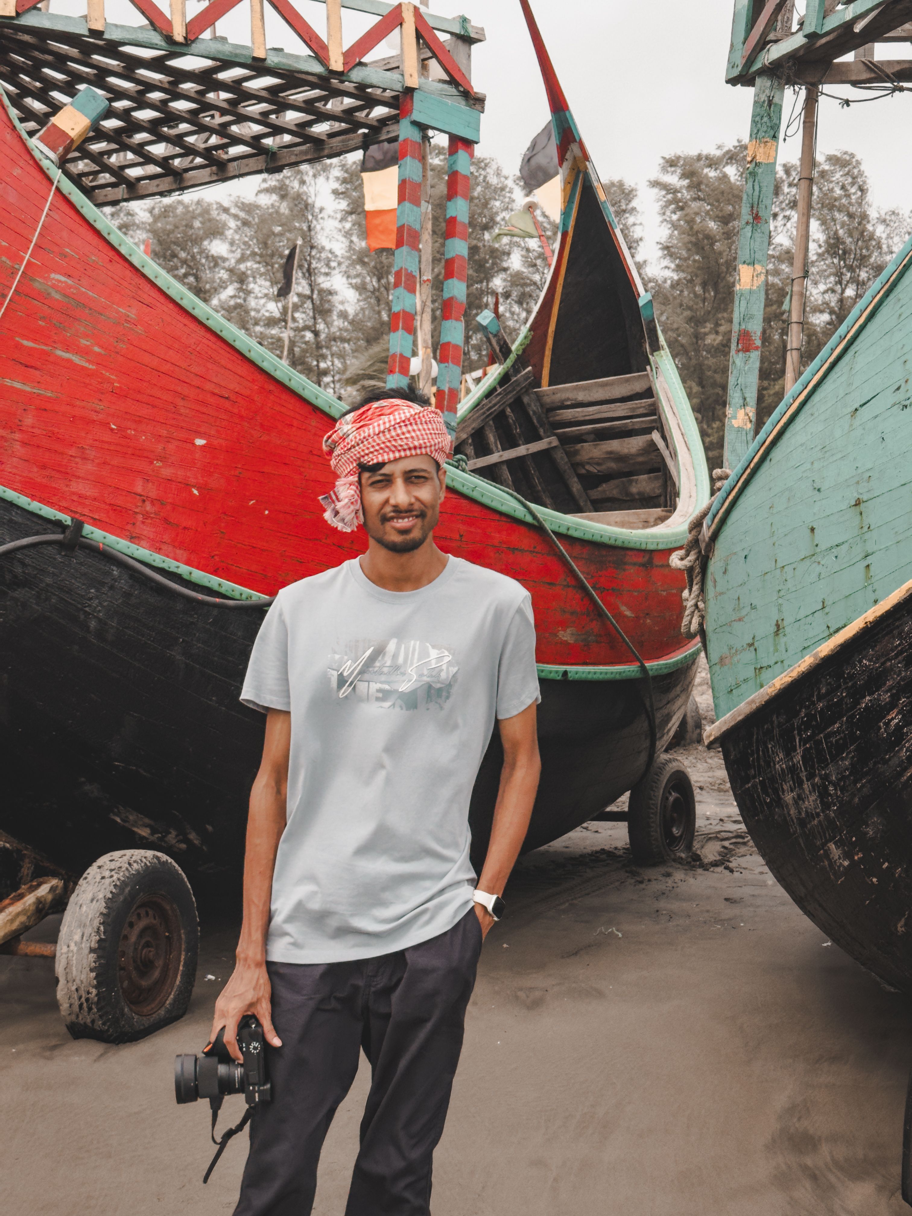 Young man with a camera stands beside vibrant boats on the beach, offering a lively and cultural scene.