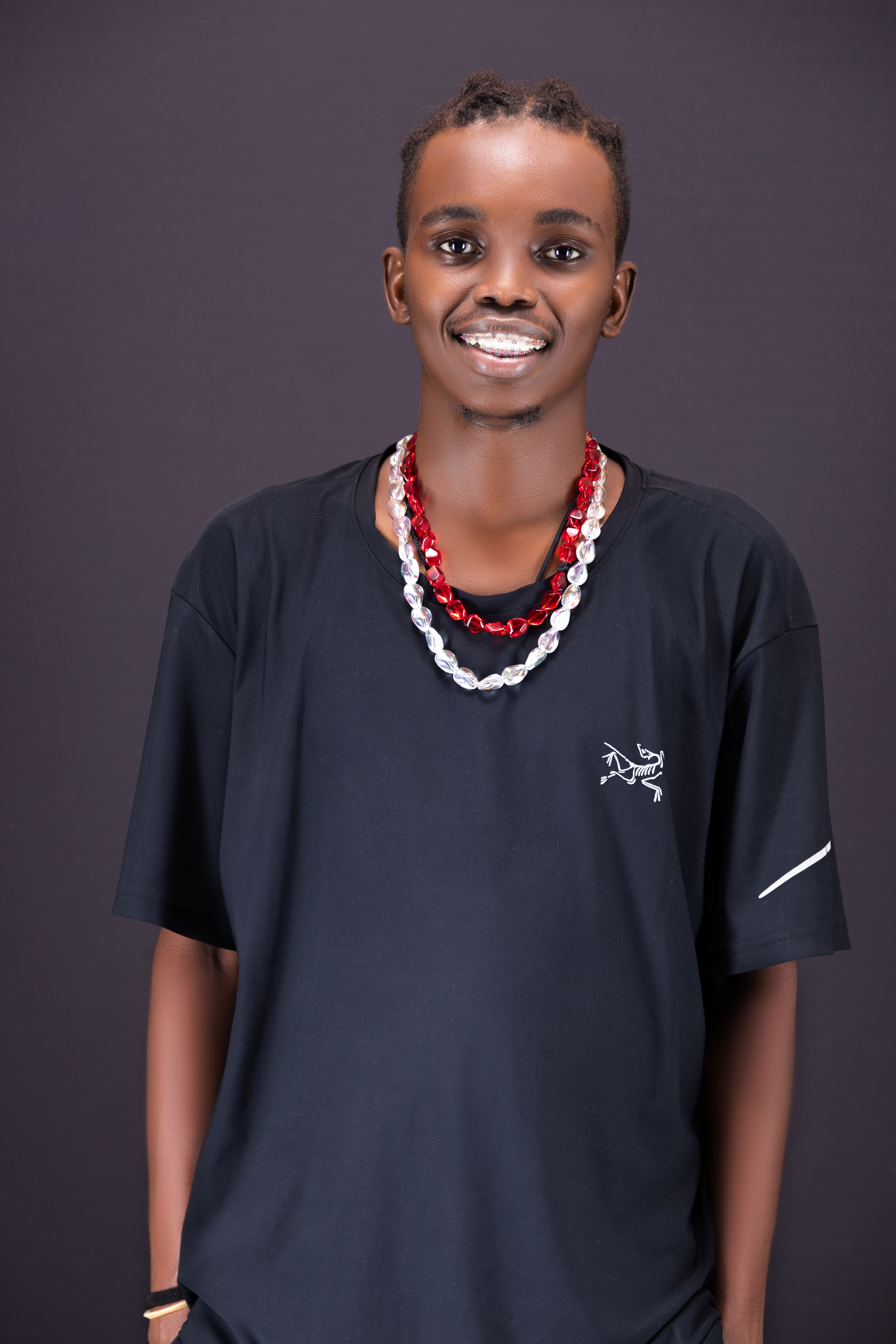 A cheerful young man wearing a black shirt and beads against a dark backdrop.