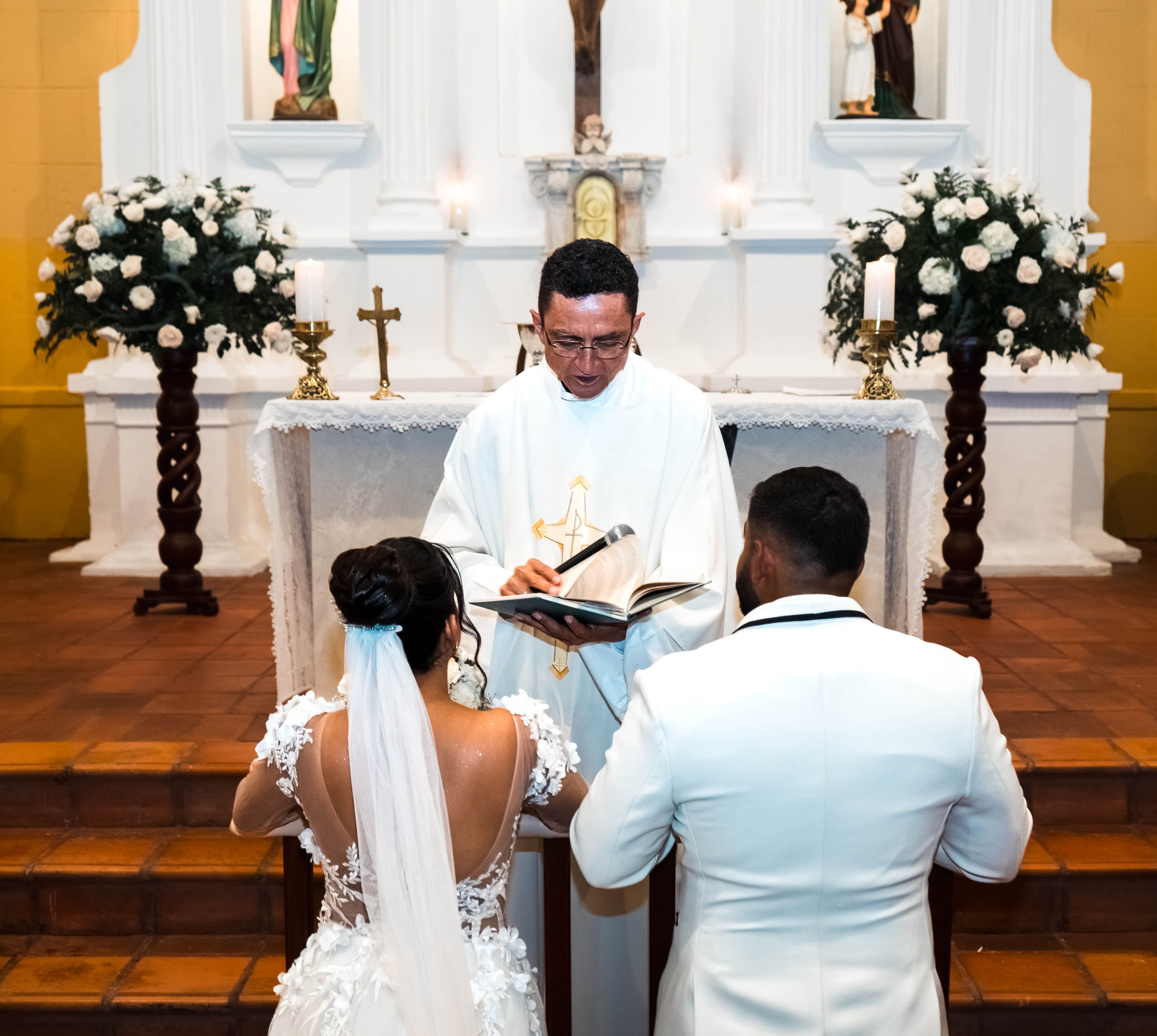 Bride and groom kneeling before priest during Catholic wedding ceremony in a church.