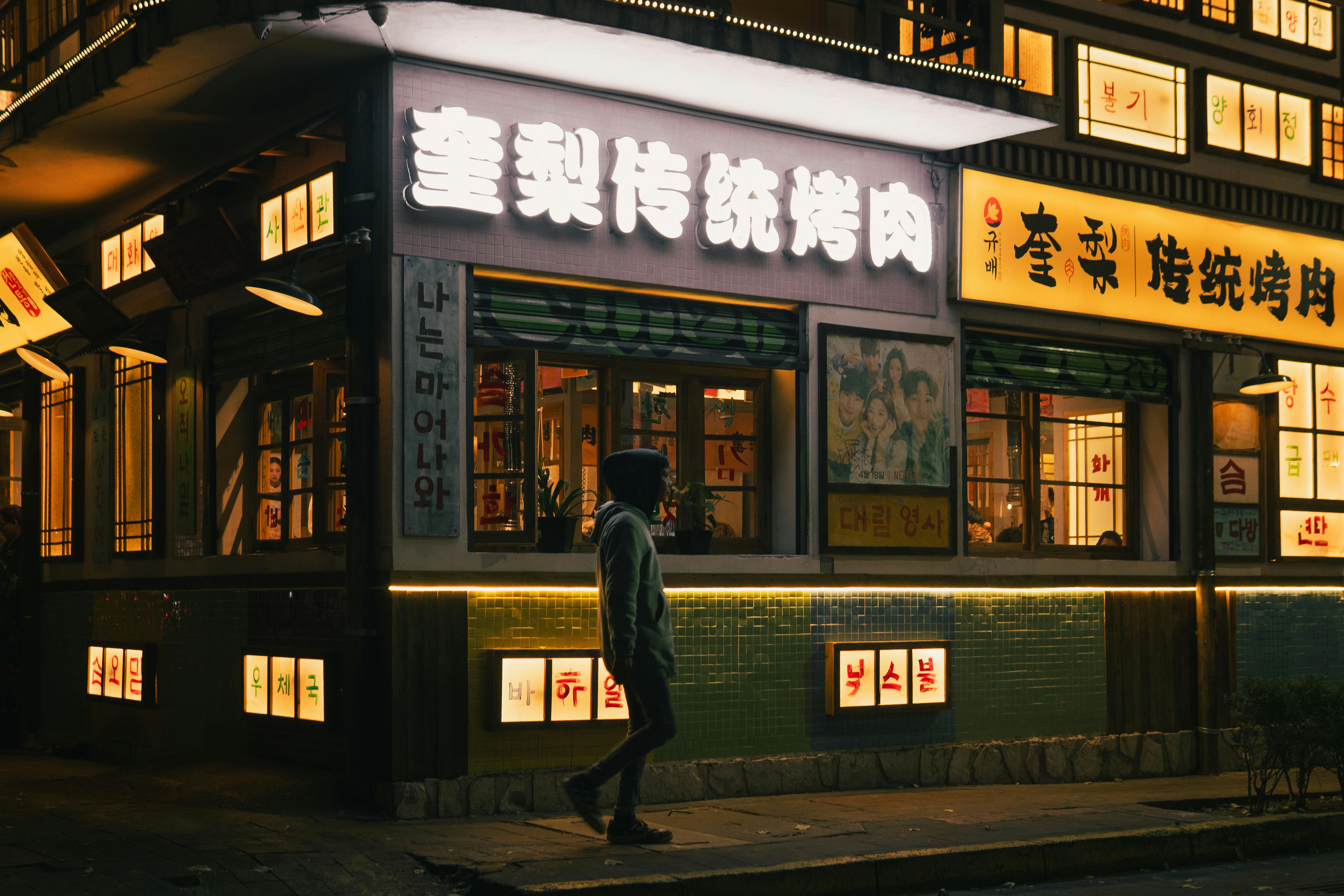 A street view of a brightly lit Korean restaurant at night, with neon signs and a lone person walking by.