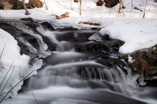 Free A tranquil winter stream flowing over icy rocks surrounded by snow-covered landscape. Stock Photo