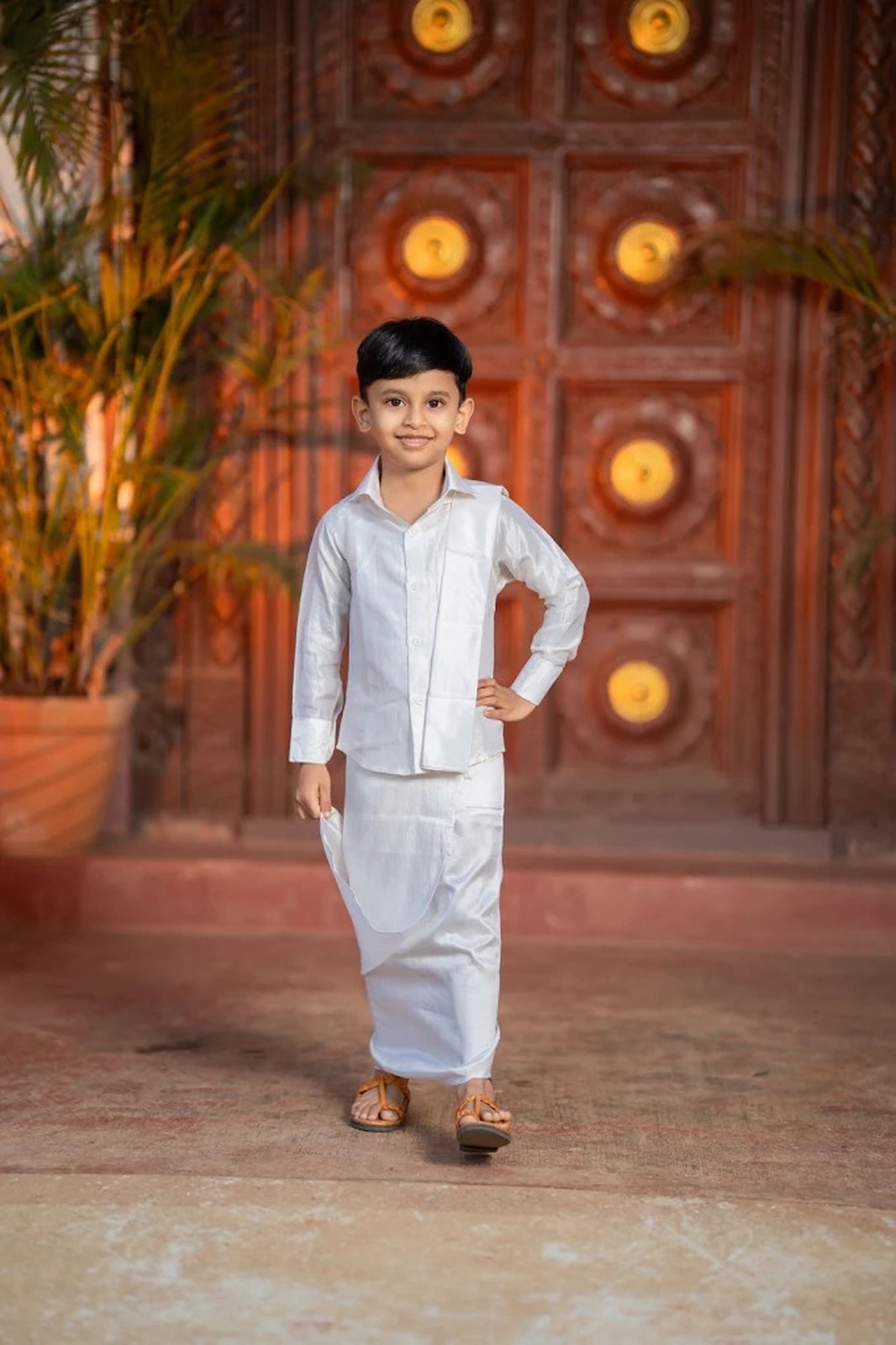 Smiling boy in traditional white attire stands before ornately carved wooden doors.